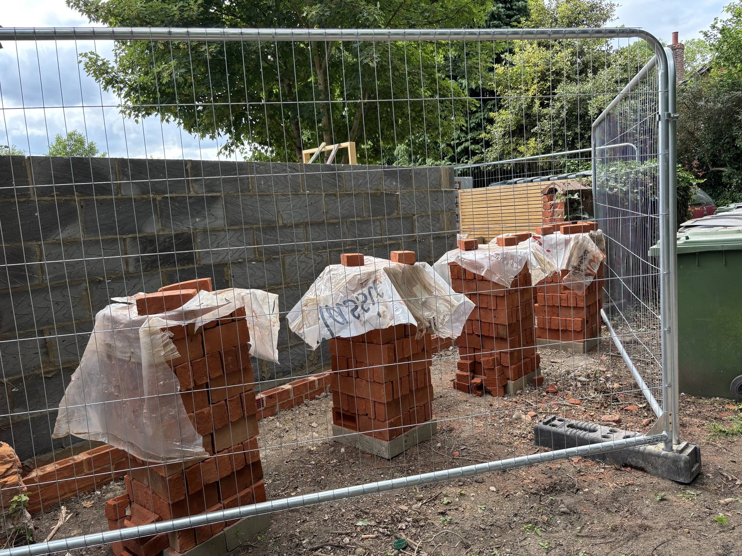 Construction site with a brick wall under construction, surrounded by a metal fence, with stacks of bricks and some construction materials on the ground, and trees in the background.