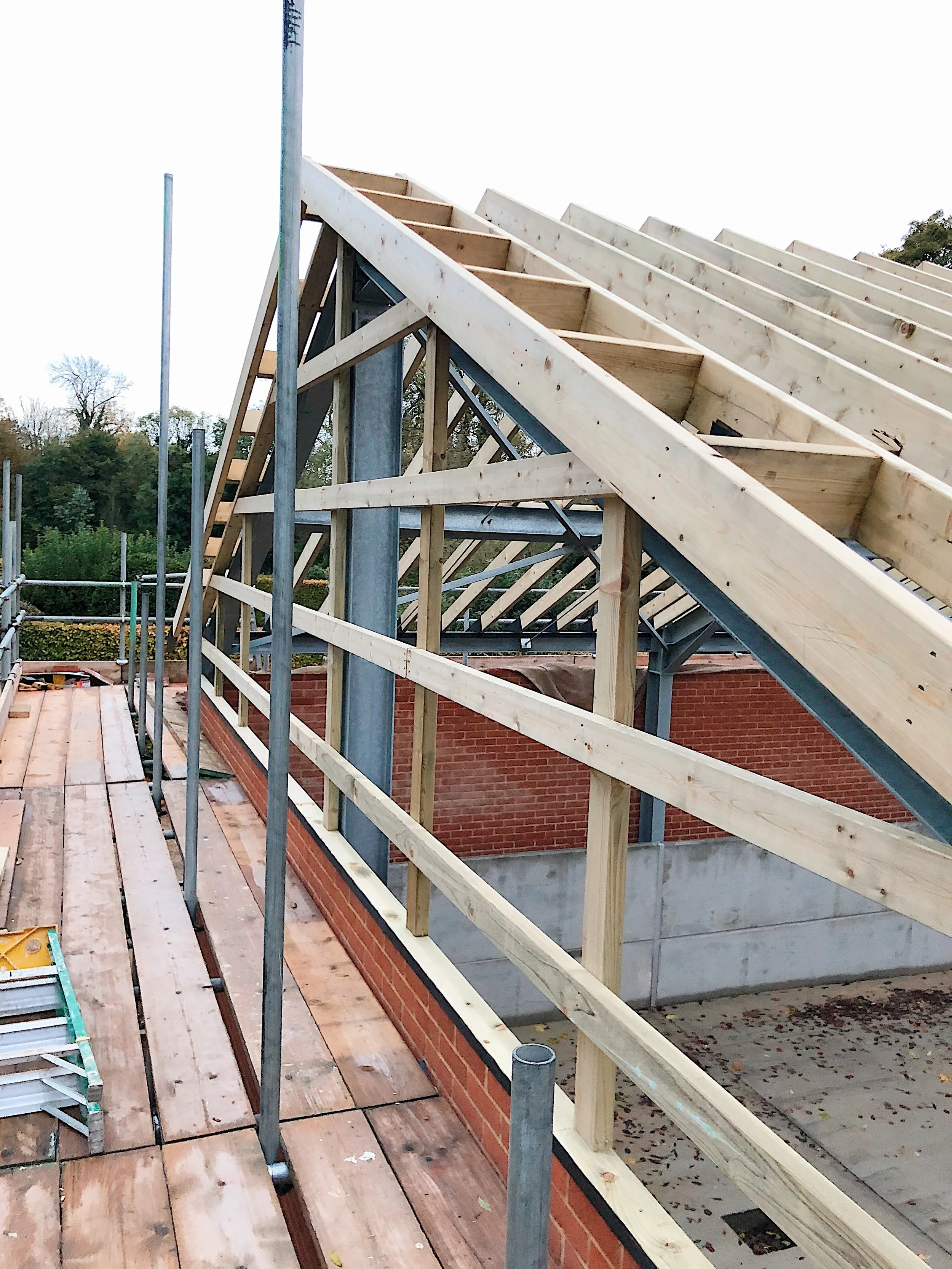 Construction site with wooden framing for a roof on top of brick walls, scaffolding and ladders present, surrounded by trees and fallen leaves.