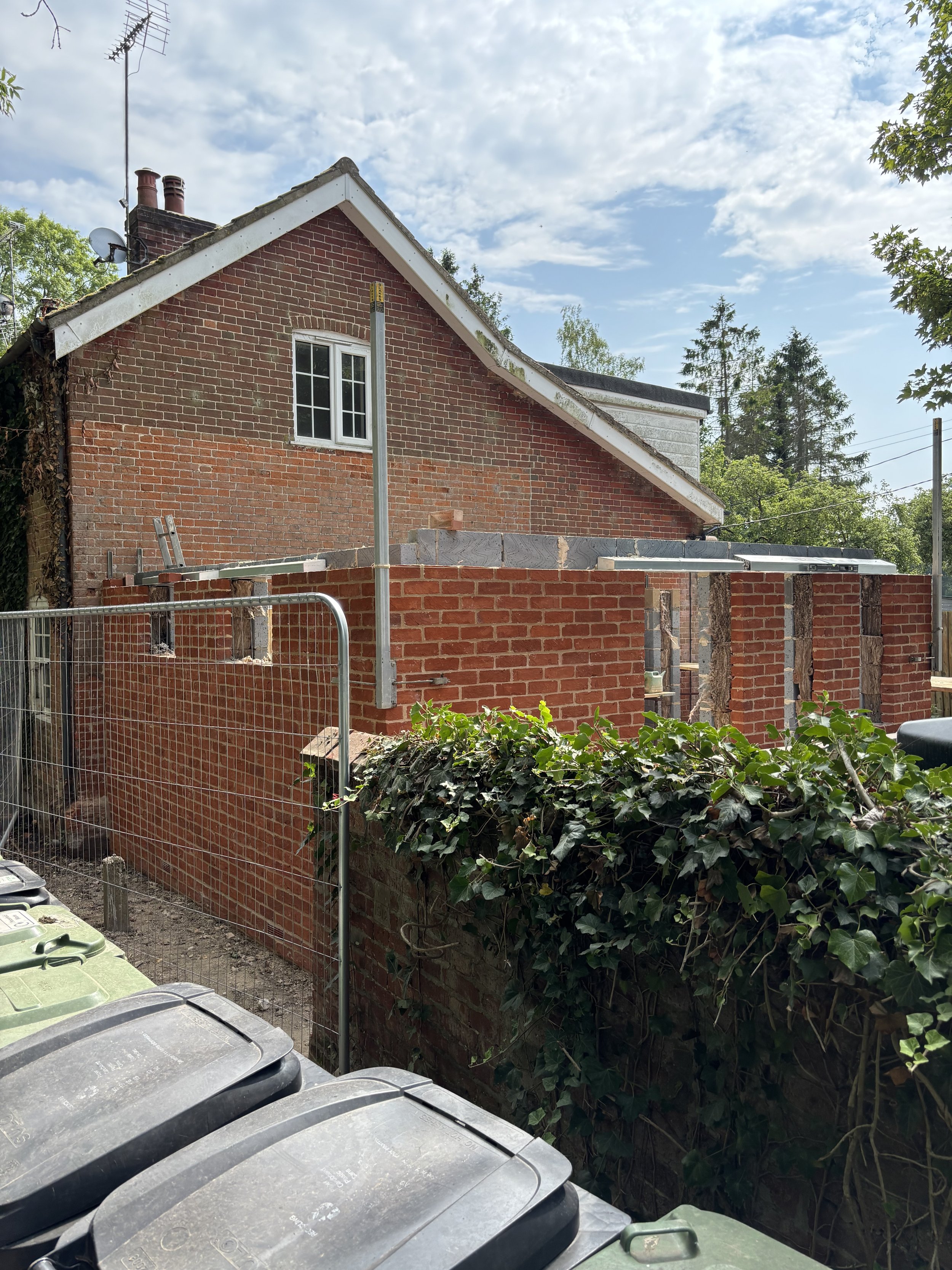 A brick house under construction with an extension being built, wooden beams supporting the new structure, a fence, green bushes, and a row of trash bins in the foreground, under a partly cloudy sky.