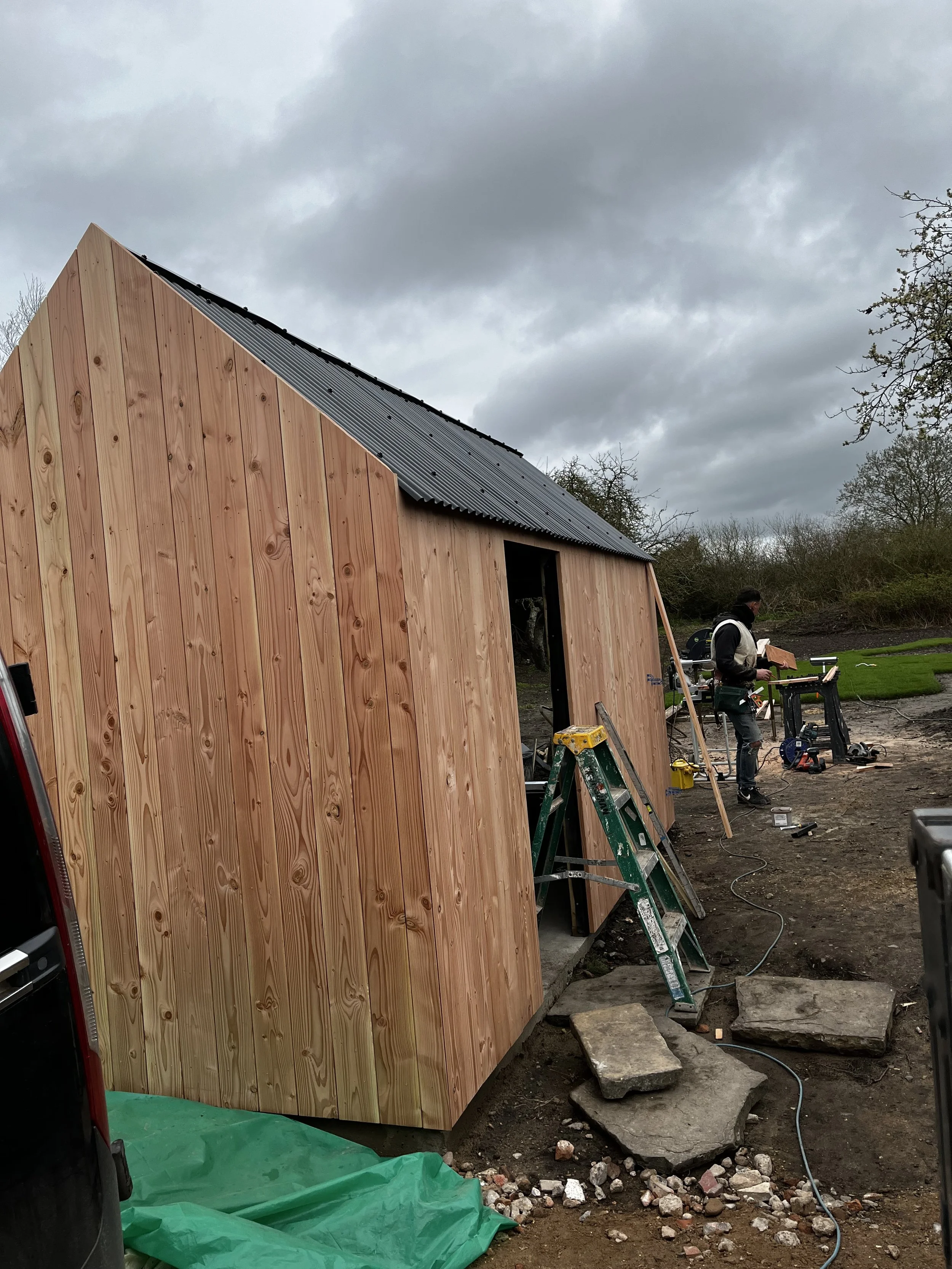 A construction site where a small wooden building with a sloped metal roof is being built. There are construction tools, a ladder, and a worker in the background working on the site, with cloudy skies overhead.