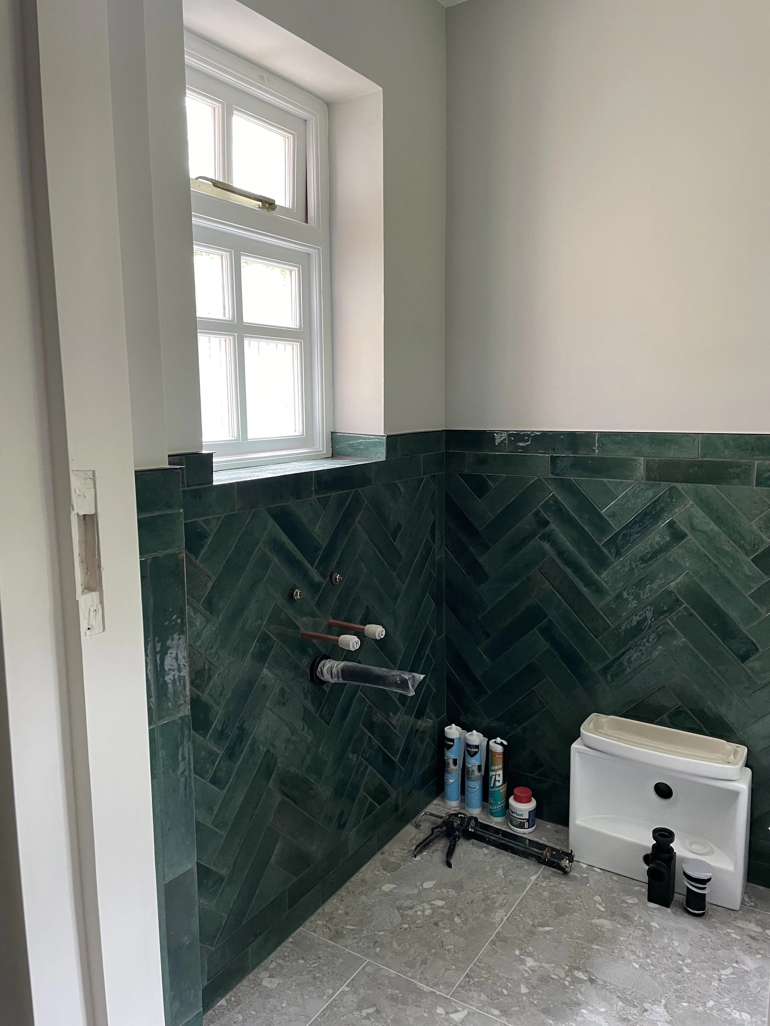 A bathroom under construction with green tile on the lower half of the wall, a window, and various tools and supplies on the floor, including cans of sealant, a small cabinet, and construction tools.