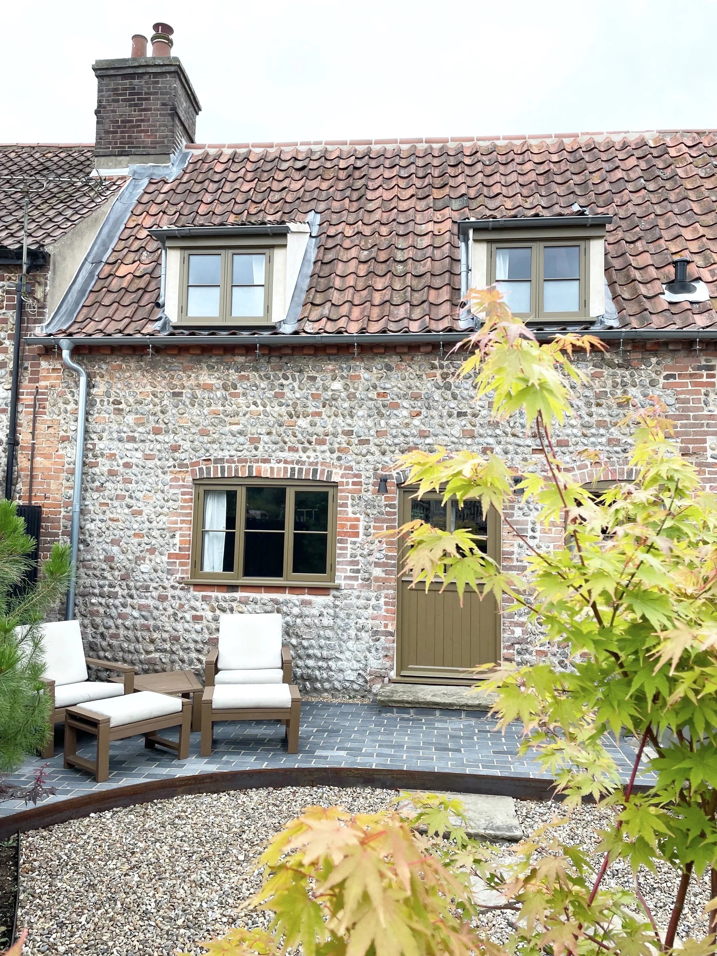 Rear view of a brick house with a stone facade, two dormer windows, outdoor seating with chairs, trees and plants in the backyard.