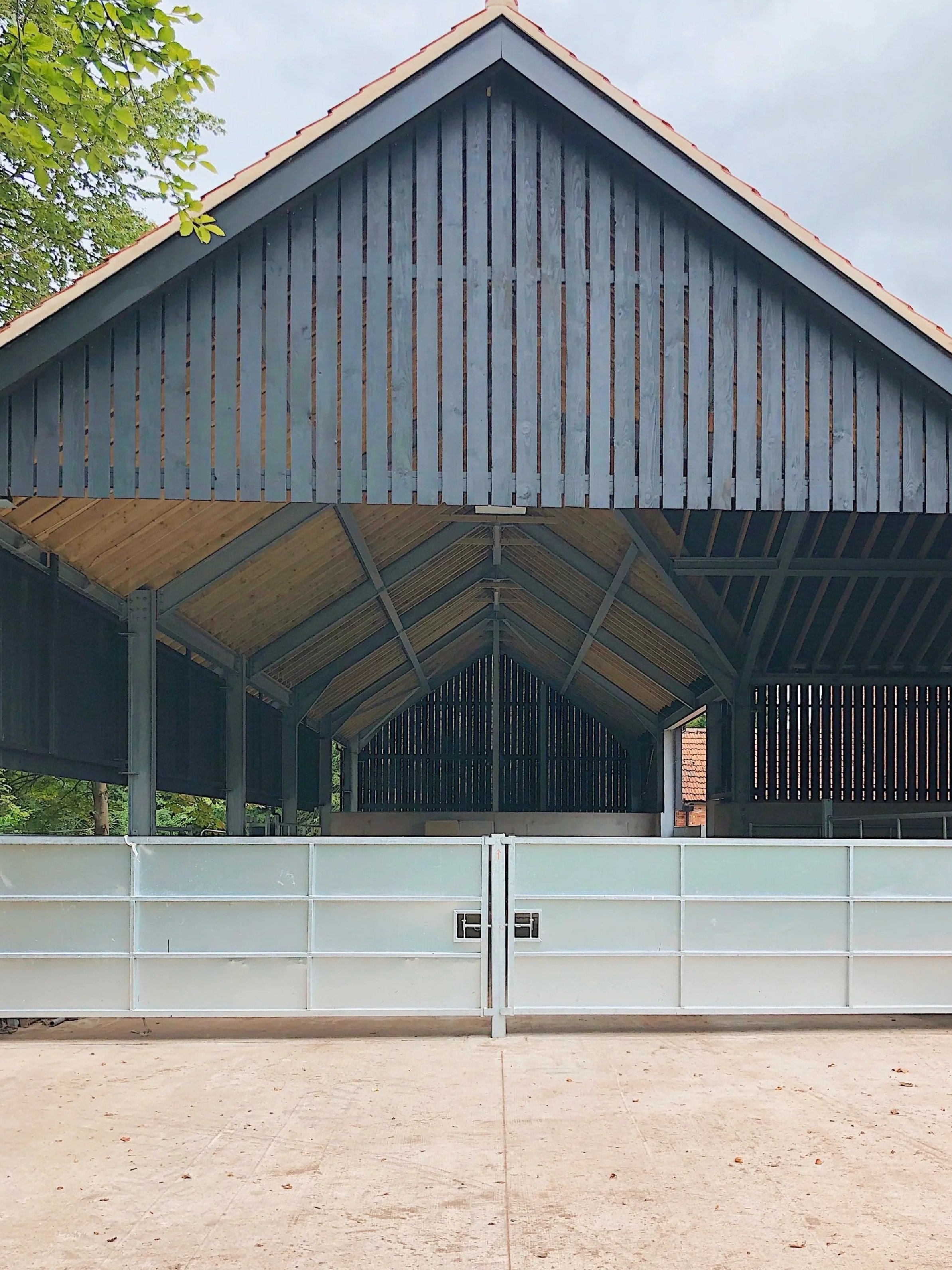 An indoor arena or barn with a high, gabled roof. The roof has wooden beams and a metal frame, and the walls are made of slatted wood panels and metal. The front of the arena has a white fence or barrier. Some trees and a building's roof are visible 