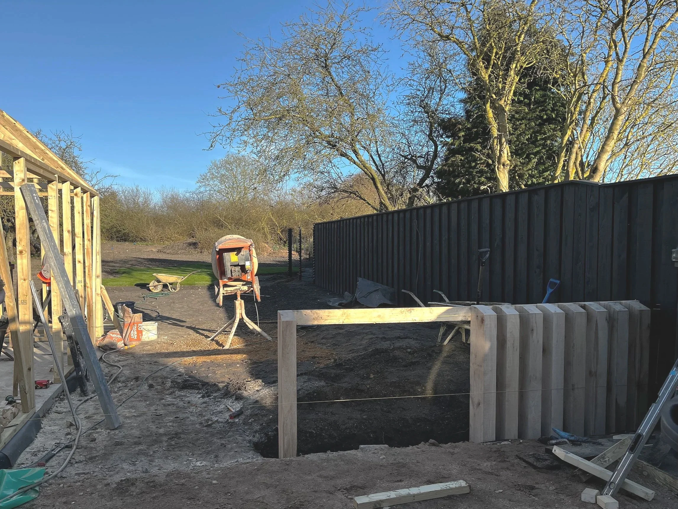 Construction site with wooden framing, metal fencing, construction tools, wet ground, and leafless trees under a clear blue sky.
