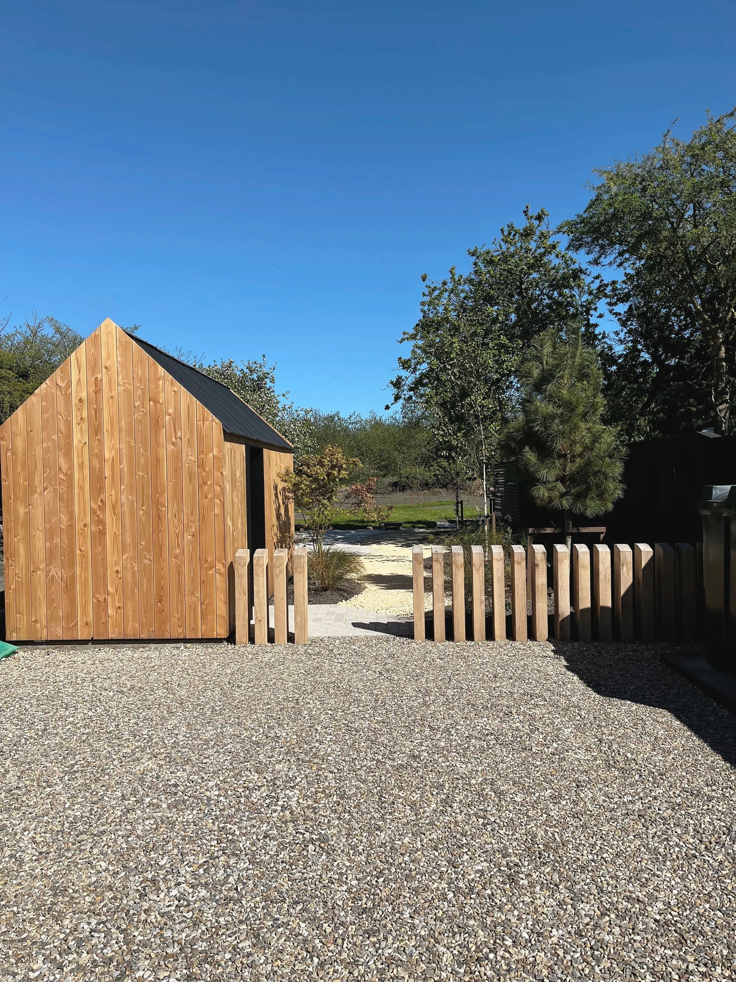 View of a backyard with a wooden shed on the left, a gravel ground, a wooden fence, and trees with green leaves against a clear blue sky.