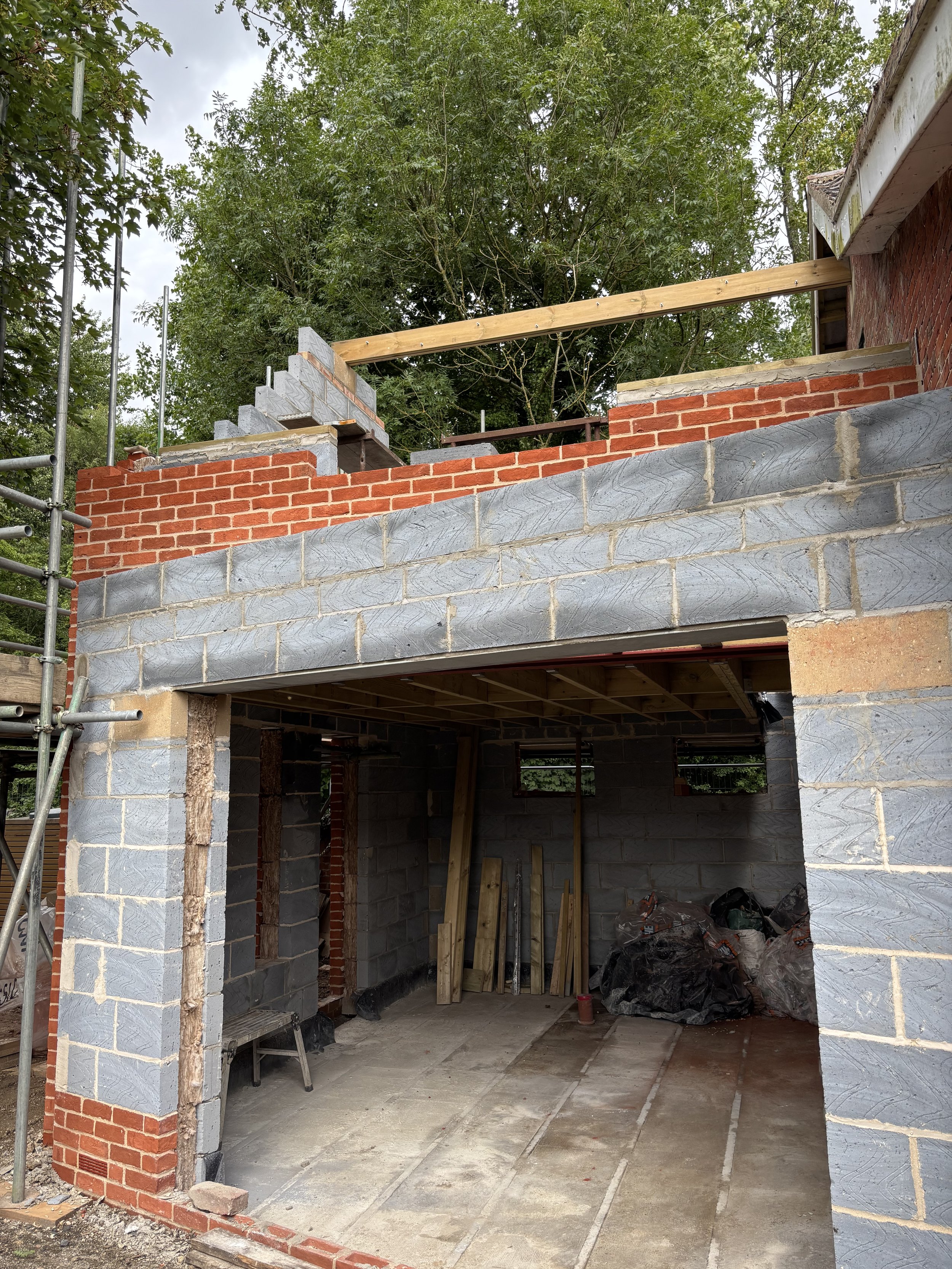 Front view of a house under construction with scaffolding on the left, brick walls, concrete blocks, and construction materials inside, with trees in the background.