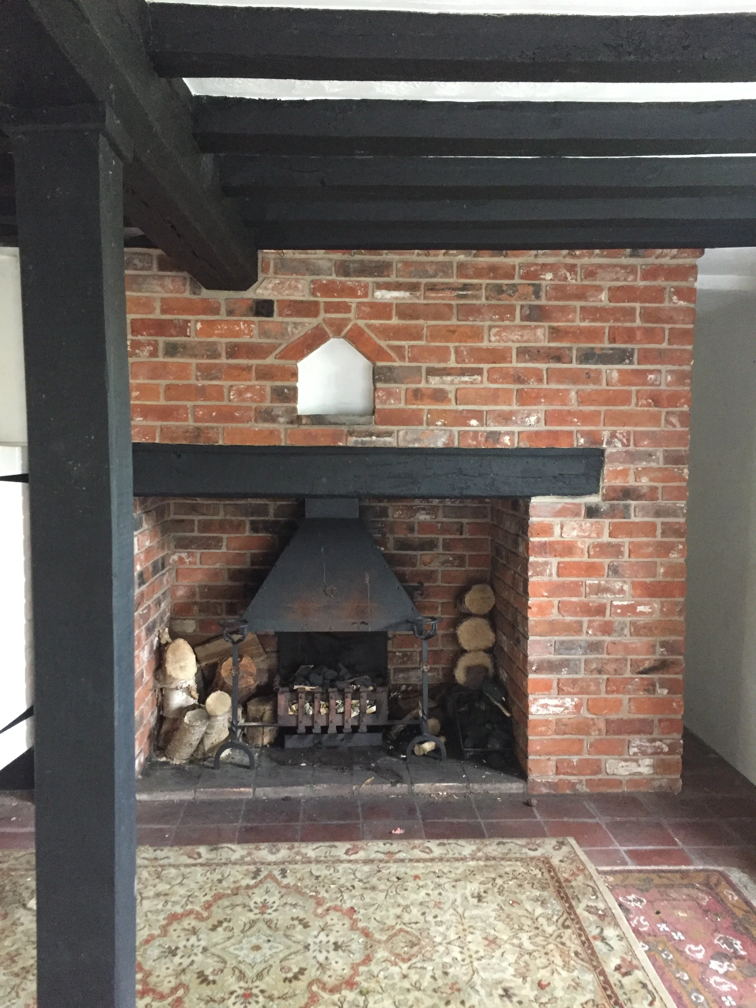A fireplace with a brick surround, a black metal hood, and a wood-storage area on the sides, situated on a tiled floor with a decorative rug partially visible in front.