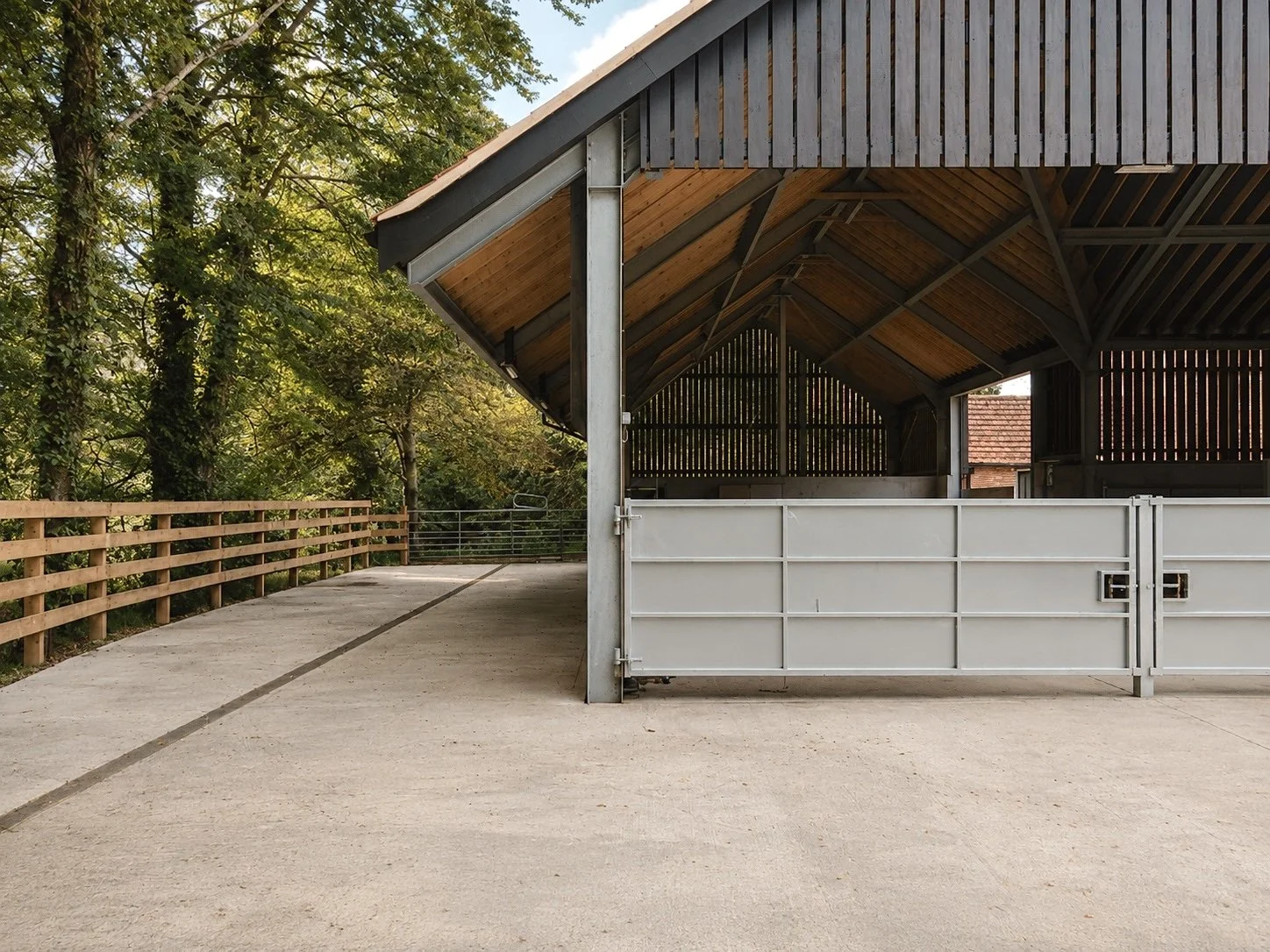 Empty covered horse stable with open gate, wooden and metal structure, surrounded by trees and countryside.