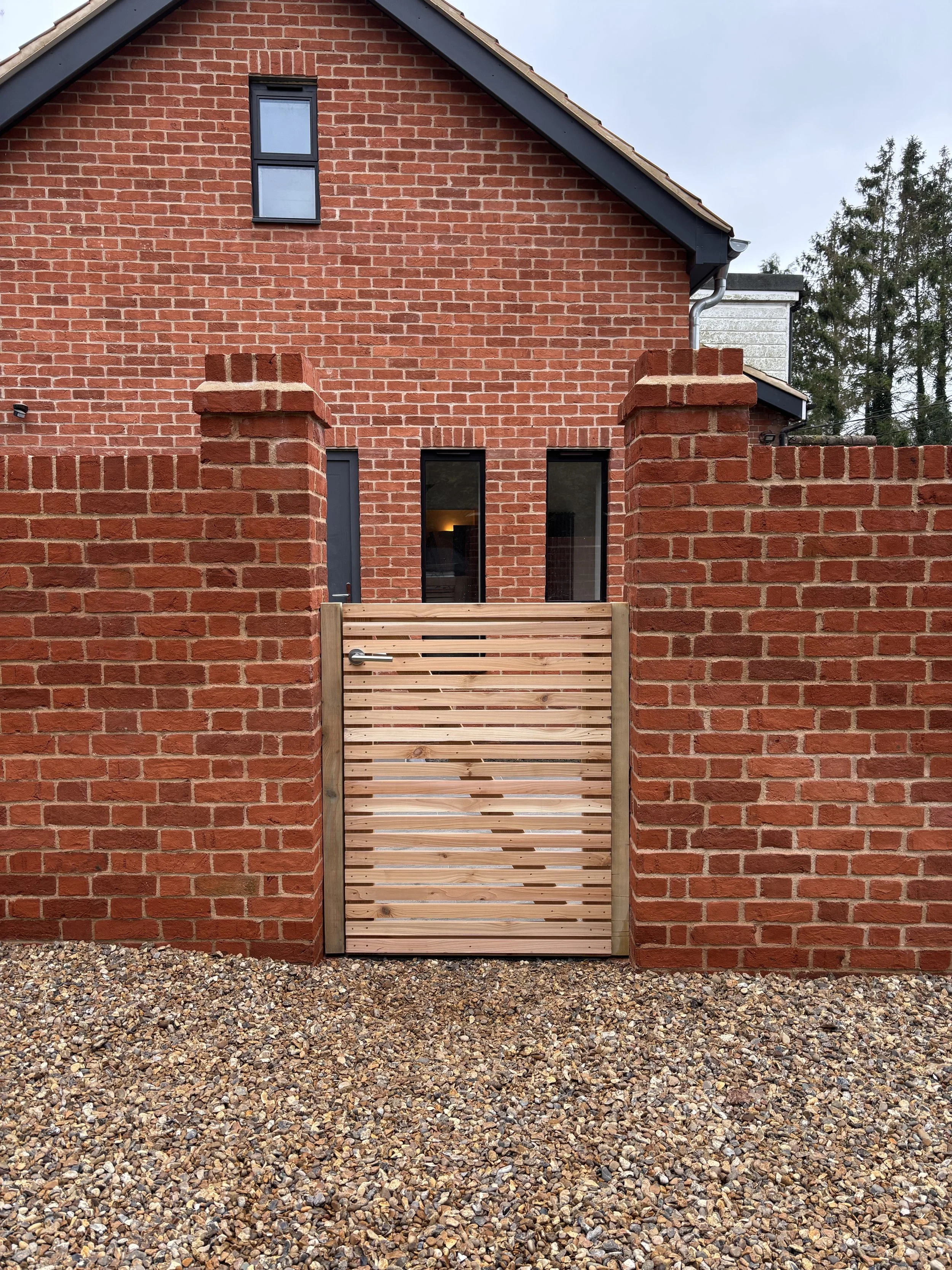 A brick house with a small black window on the upper level and two larger black windows on the lower level. In front, there's a wooden gate with horizontal slats and a metal handle, surrounded by a low brick wall. The ground is covered with small gra