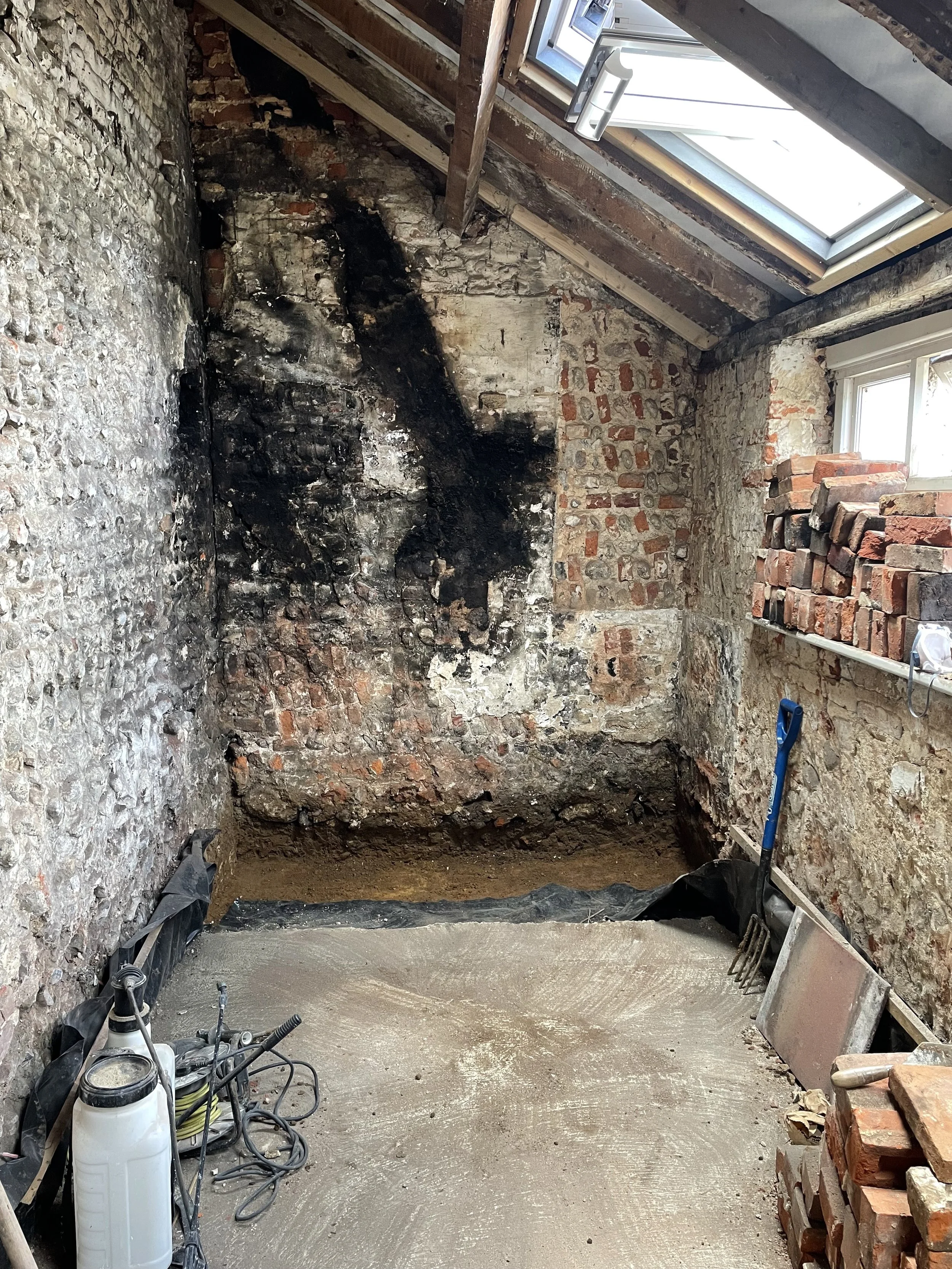 Attic renovation in progress with exposed brick and stone walls, a large window, and construction tools, including a bleach container, a trowel, a rake, and a pile of bricks.