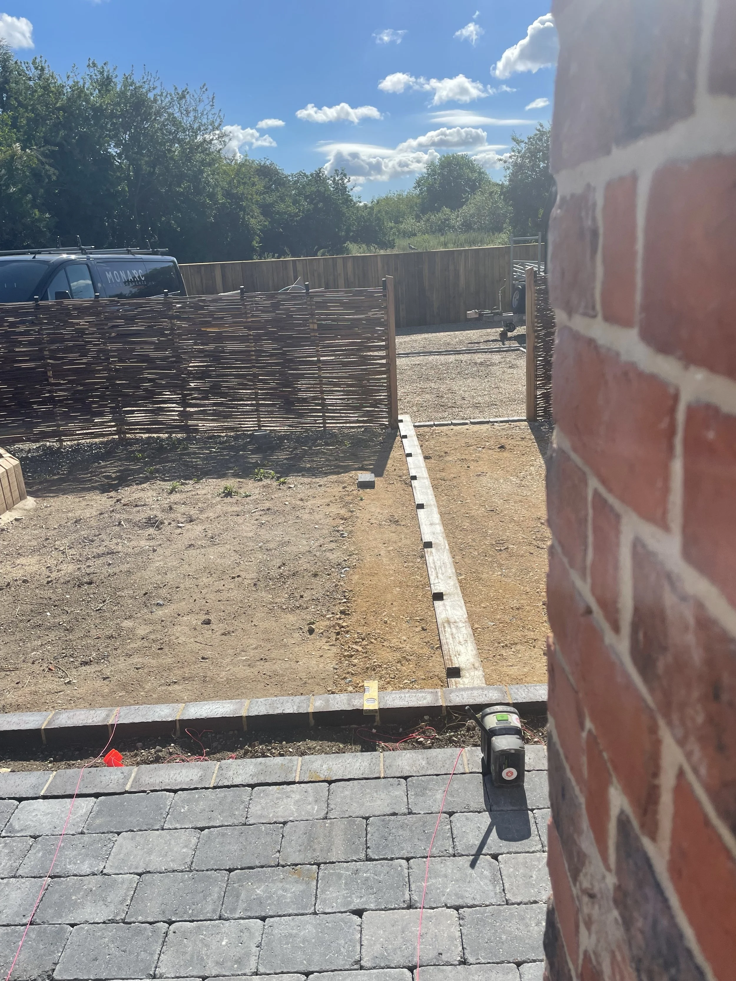 A partially constructed outdoor patio area with a row of gray stone pavers, some red string line, and building materials. In the background, there's a grassy yard, a wooden privacy fence, a black van with the word 'MONARCH' on the side, a trailer, tr
