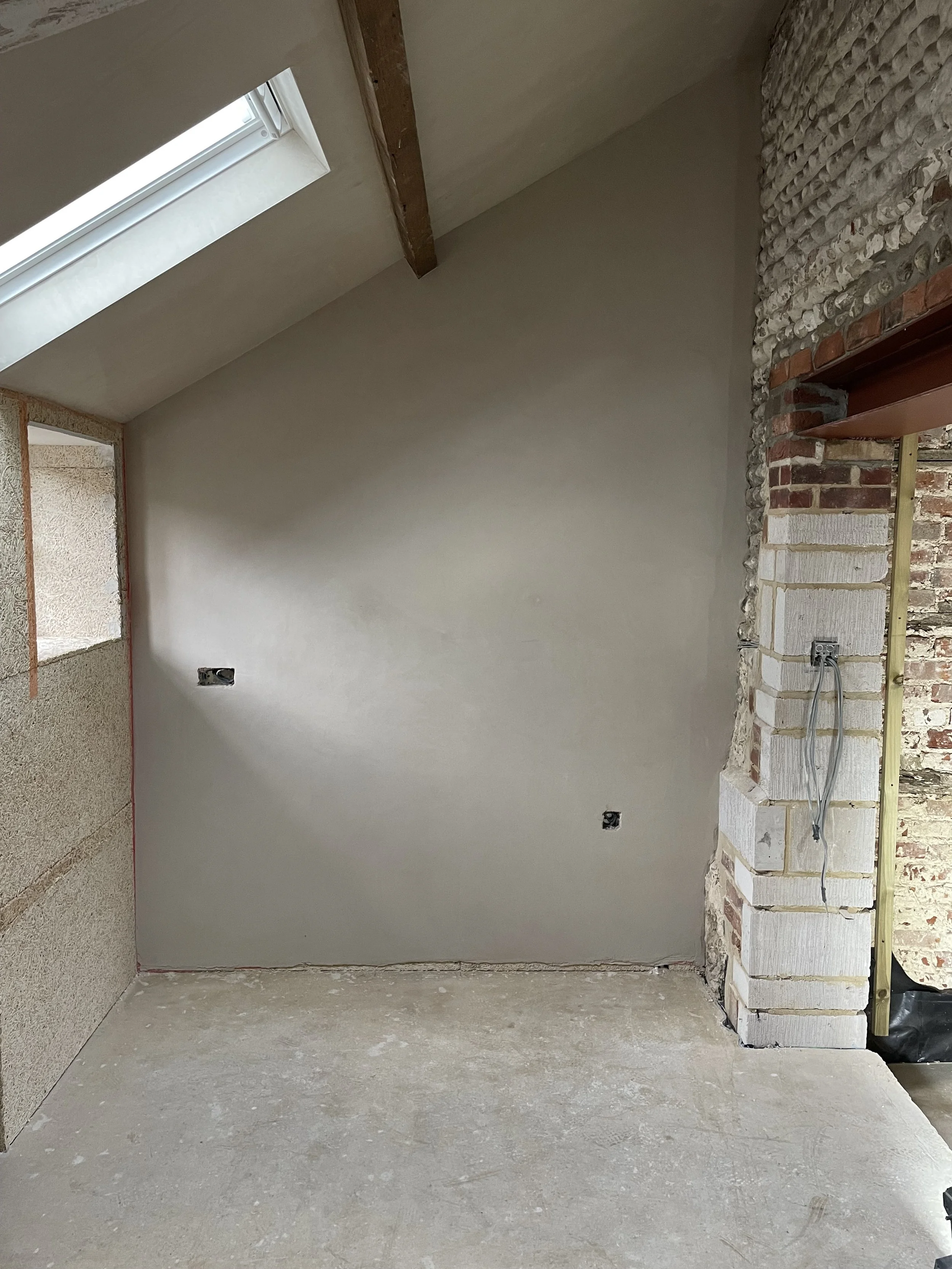 Interior of a construction site featuring a partially finished room with exposed brick and stone wall, drywall, an electrical outlet with wires, and a skylight window in the ceiling.