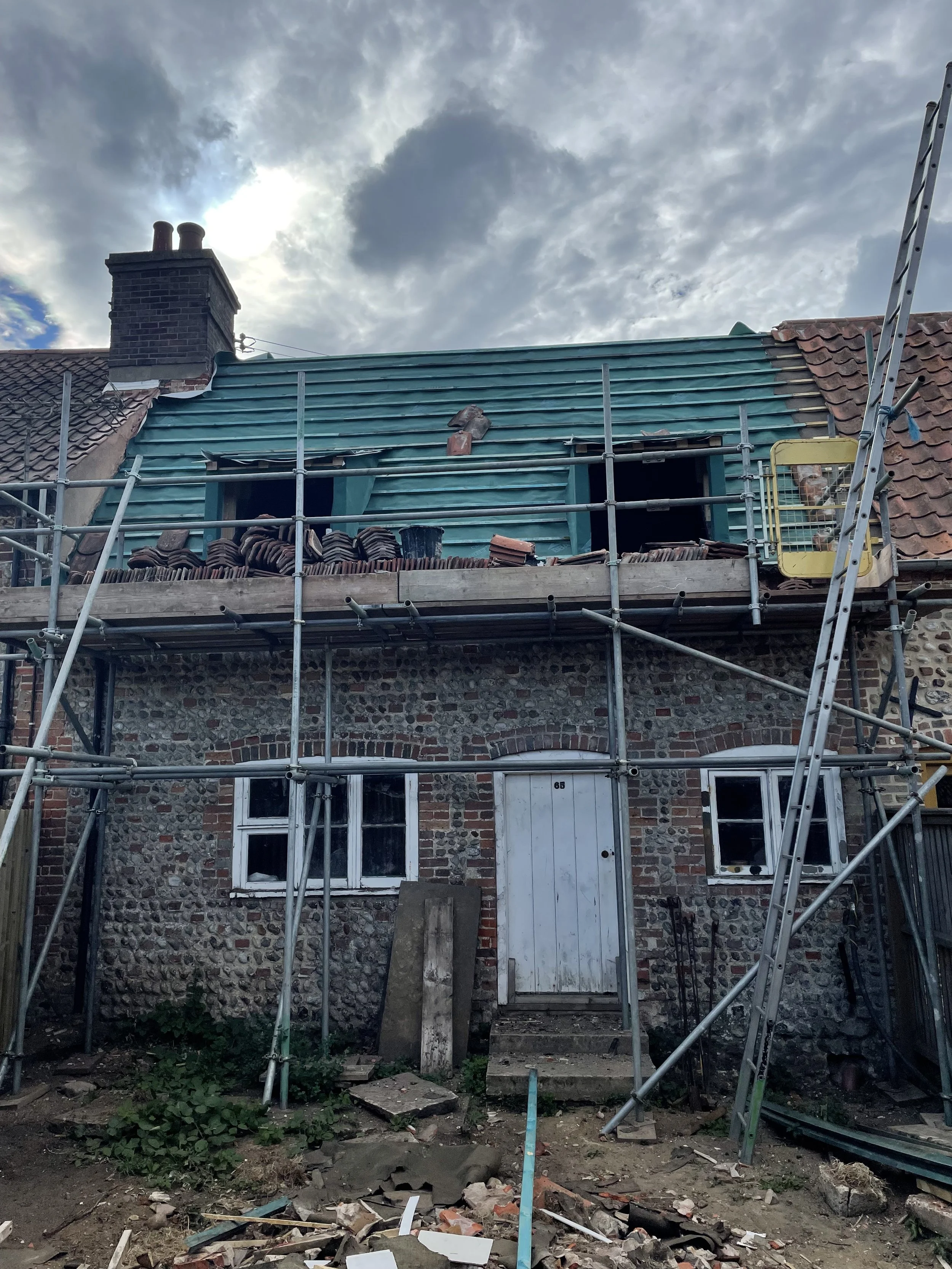The image shows a house under construction or renovation, with scaffolding around the building. The roof is partly covered with new green metal roofing sheets, and some bricks are visible on the roof. The house has a stone and brick exterior with two