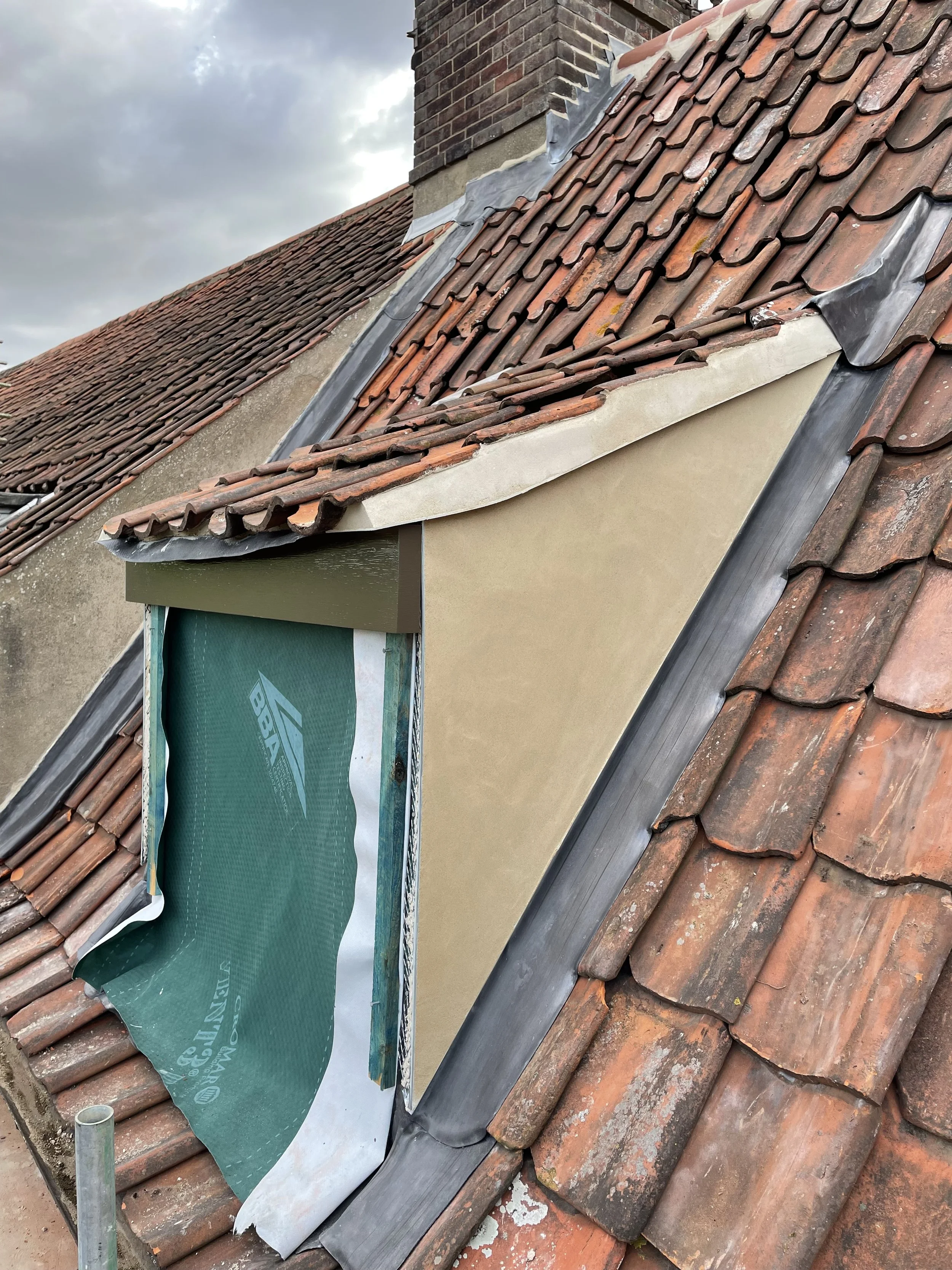 Construction workers repairing a roof with terracotta tiles, insulation material, and flashing, under a cloudy sky.