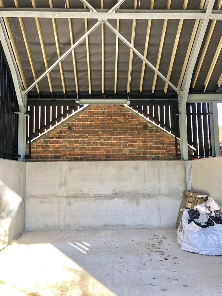 View of an unfinished outdoor structure with a concrete floor and wall, brick wall in the background, and a roof with wooden support beams. A pile of trash and wooden pallets is in the corner.