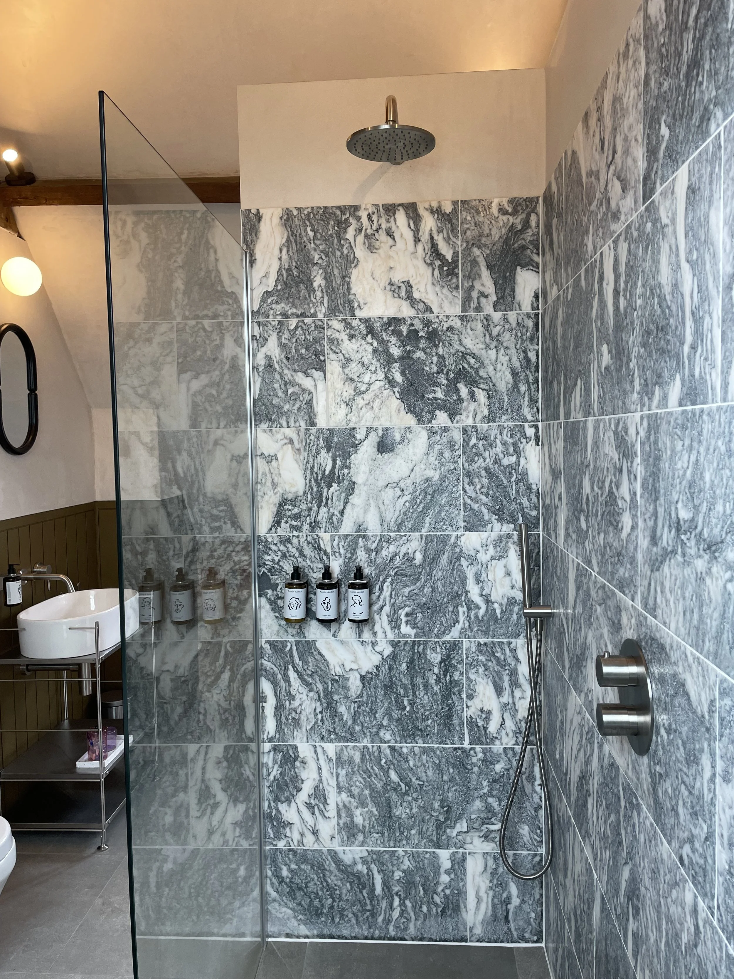 Modern bathroom shower with black and white marble tiles, a round rainfall showerhead, a handheld shower, and bottles of shampoo and soap. A sink and mirror are visible on the left.