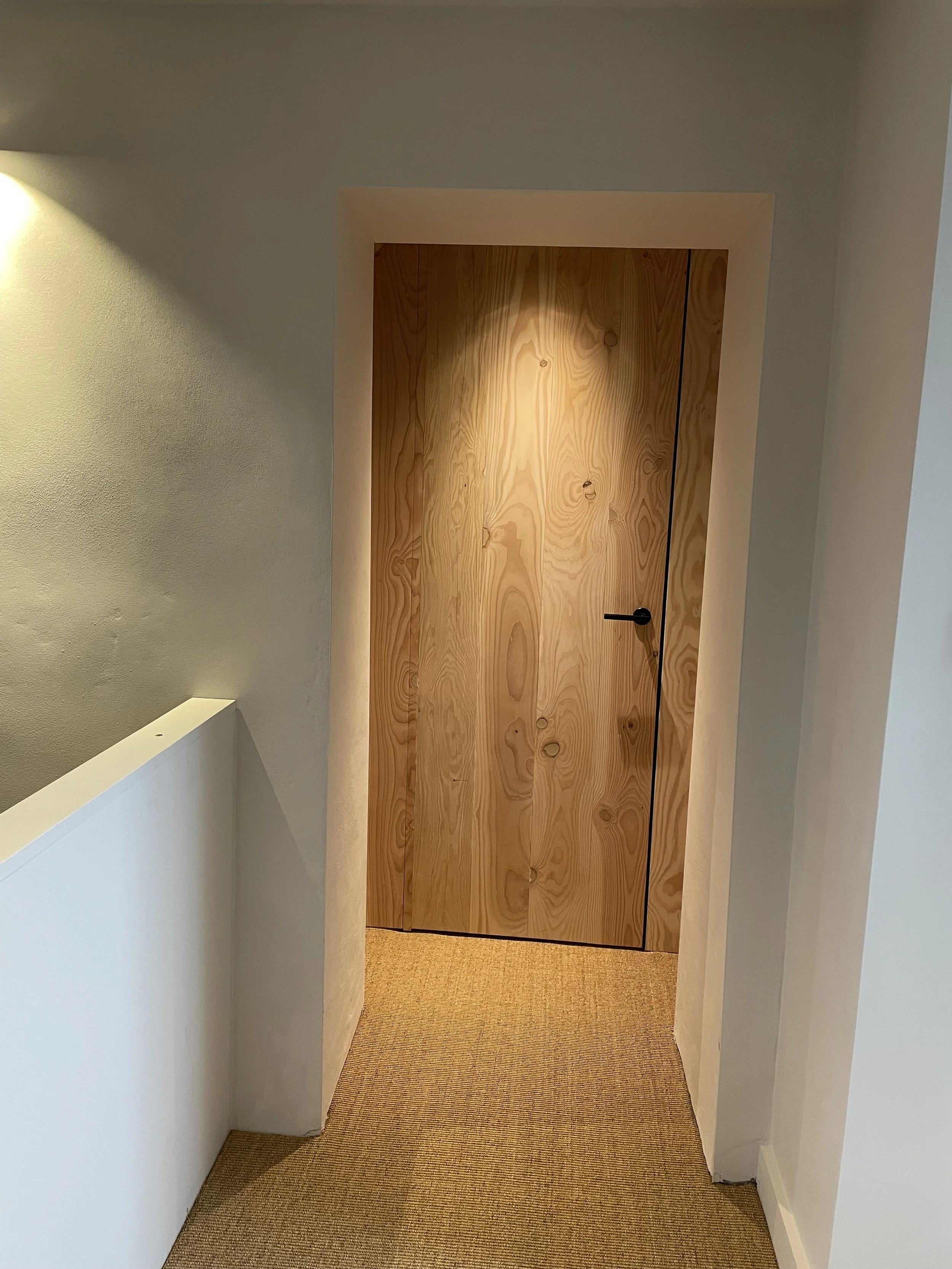 An interior hallway with a wooden door in the center, surrounded by white walls and a beige carpeted floor.
