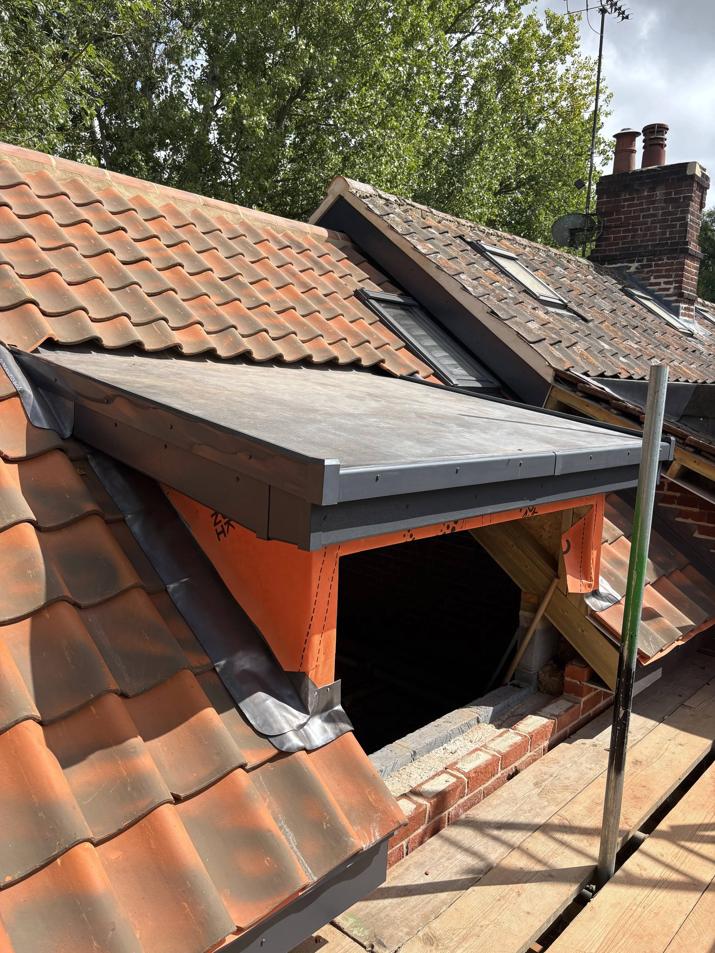 Roof under construction with tiles and several roof windows, brick chimney, and satellite dish, with green trees in the background.