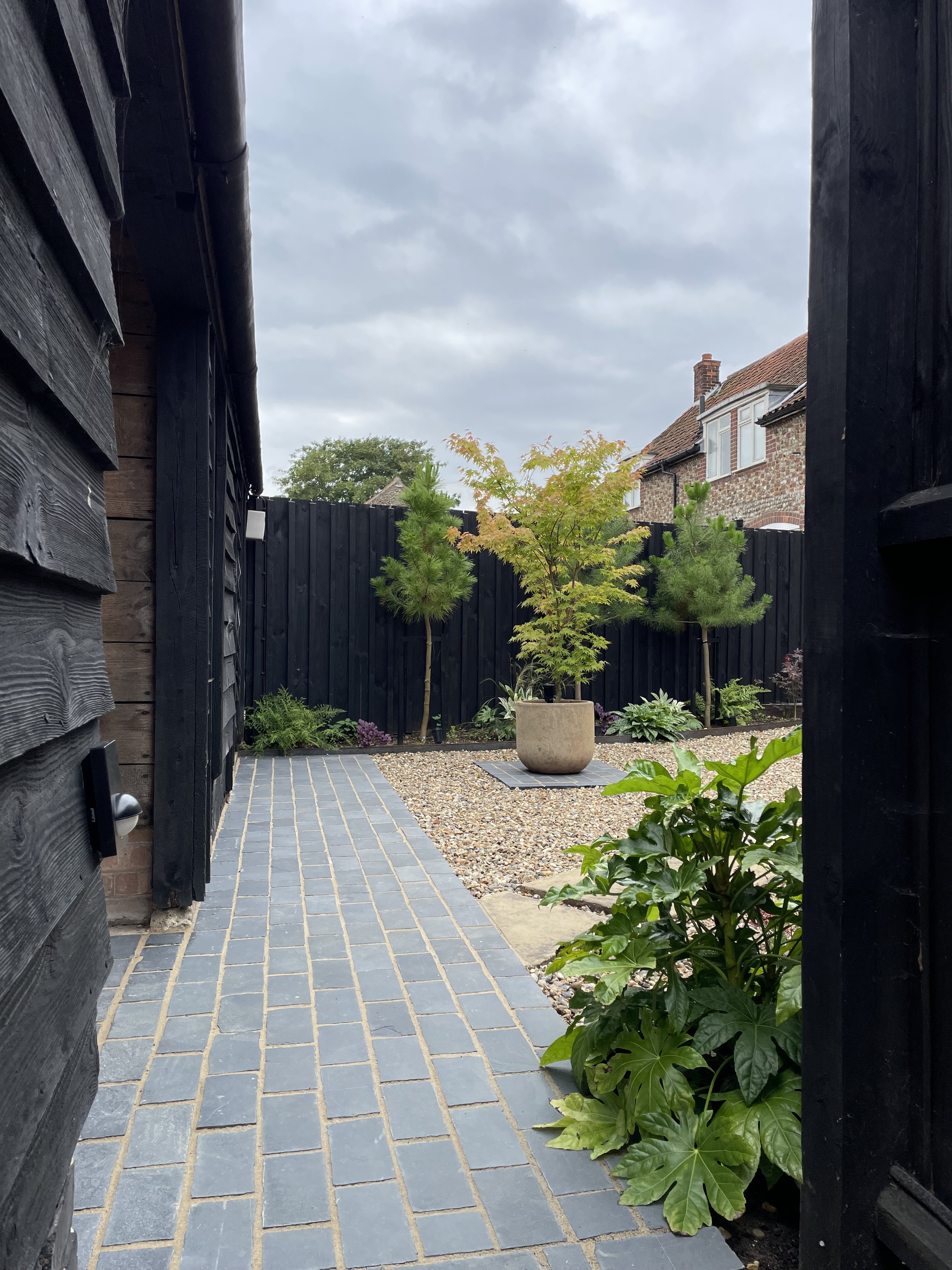 View of a backyard garden seen from between two black wooden gate posts, with a gray tiled pathway, green plants, a small tree in a pot, and a gravel bed, with a brick house and cloudy sky in the background.