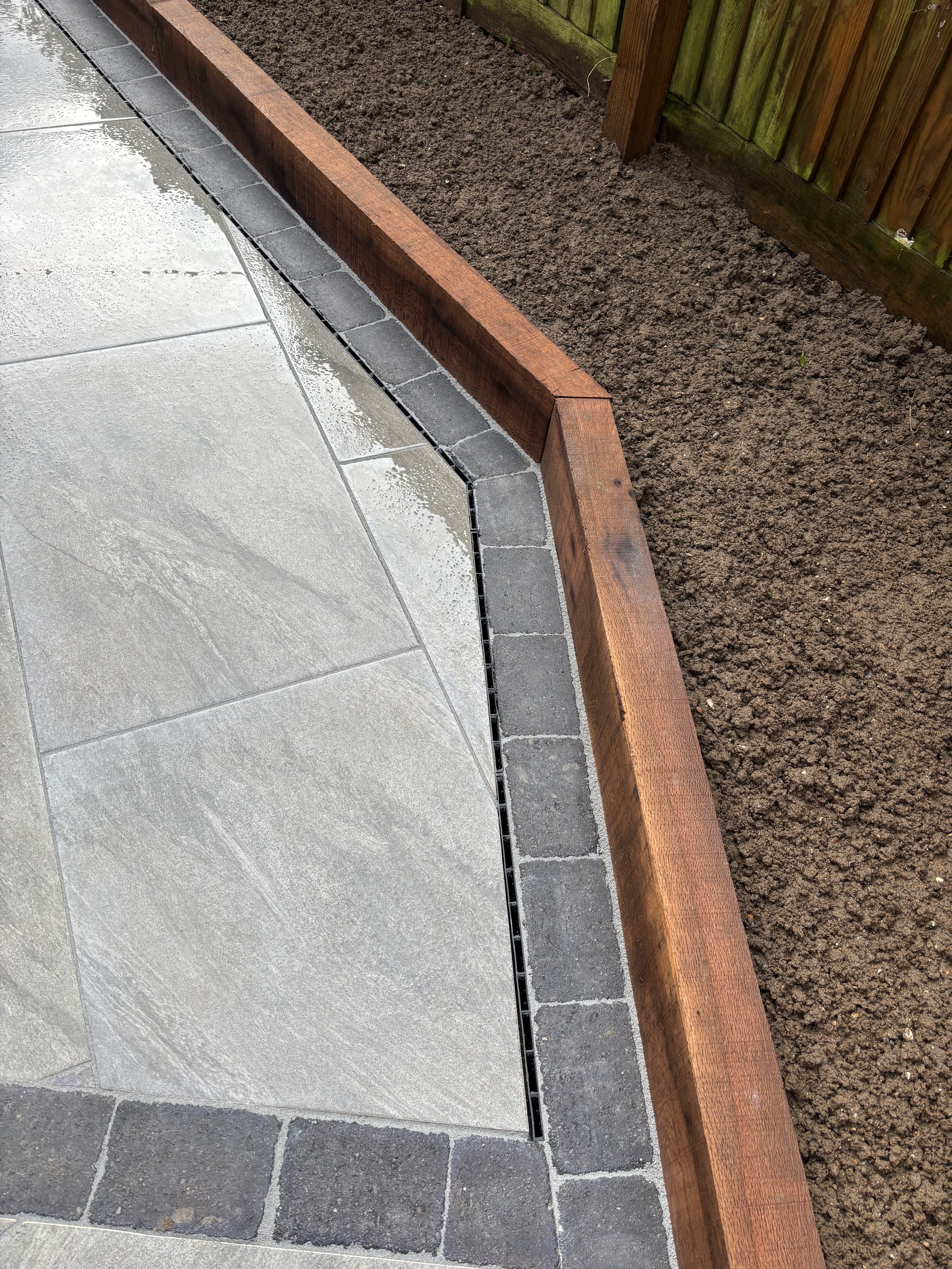 A garden bed with a wooden border and soil, with a section of gray paving stones and a drainage grate on the ground.