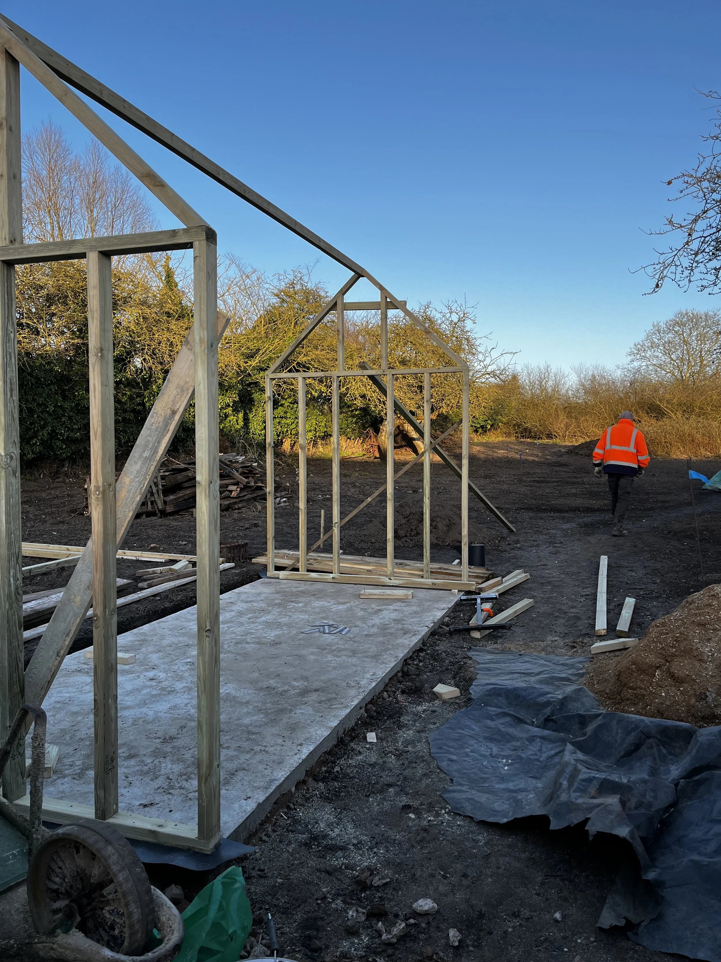 Construction site with a wooden frame structure, partially built, on concrete foundation, and a worker in an orange safety vest walking away, trees and blue sky in the background.