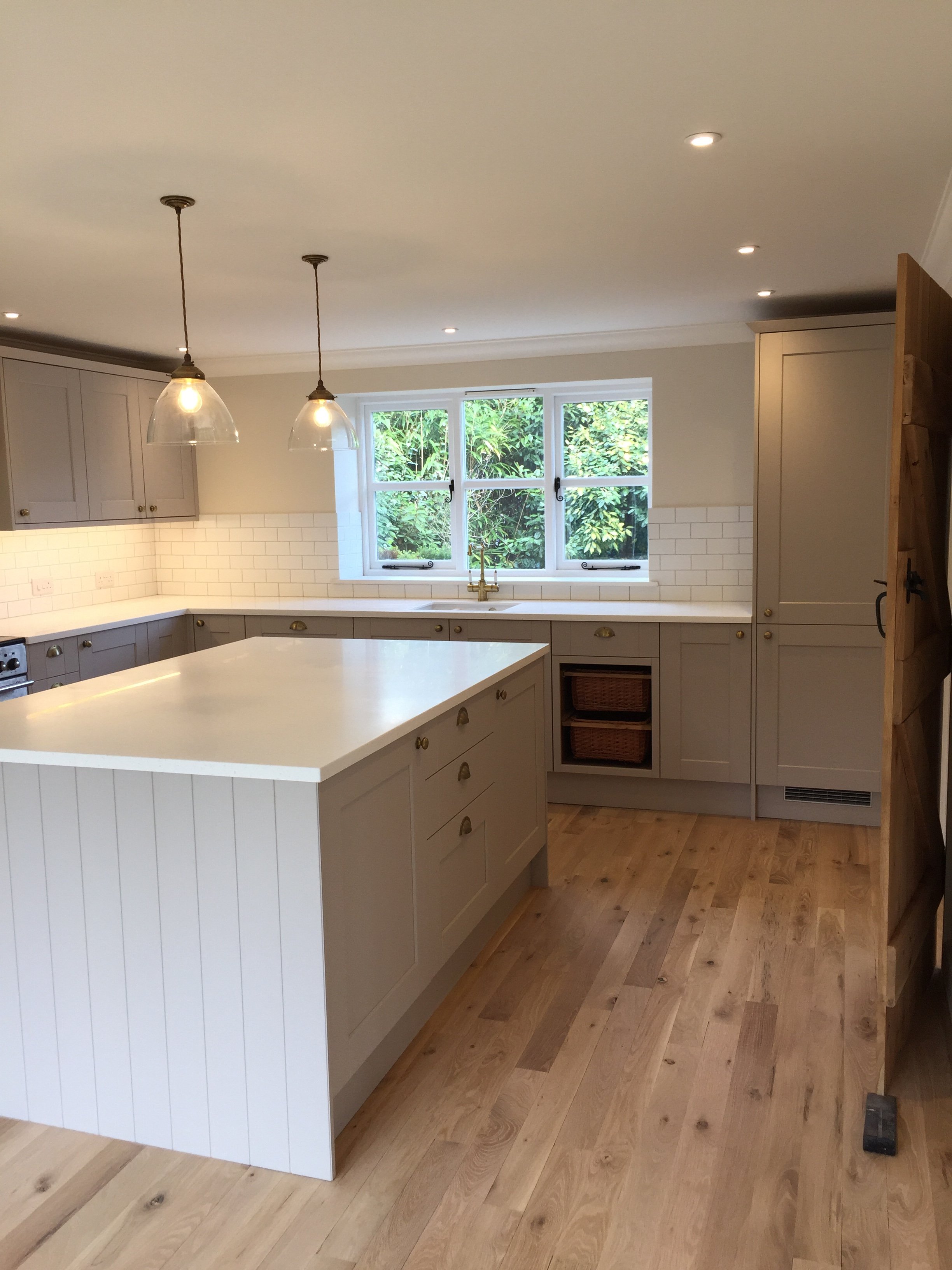 Modern kitchen with wooden flooring, light gray cabinets, white countertop, two pendant lights, and a window with greenery outside.