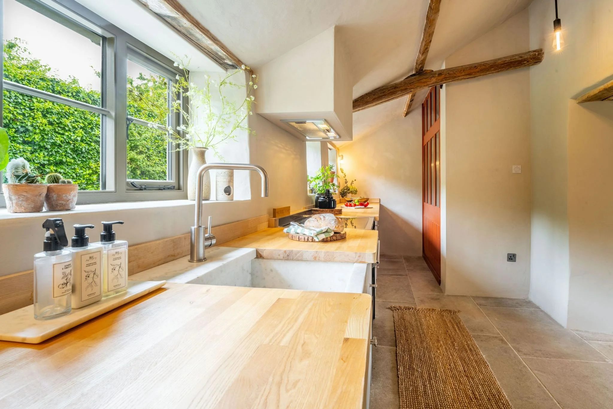 A kitchen with a large window that overlooks green trees. The countertop has a wooden surface with a sink. There are three bottles of soap or lotion near the sink. The kitchen has exposed wooden beams and warm lighting, with various plants and decora