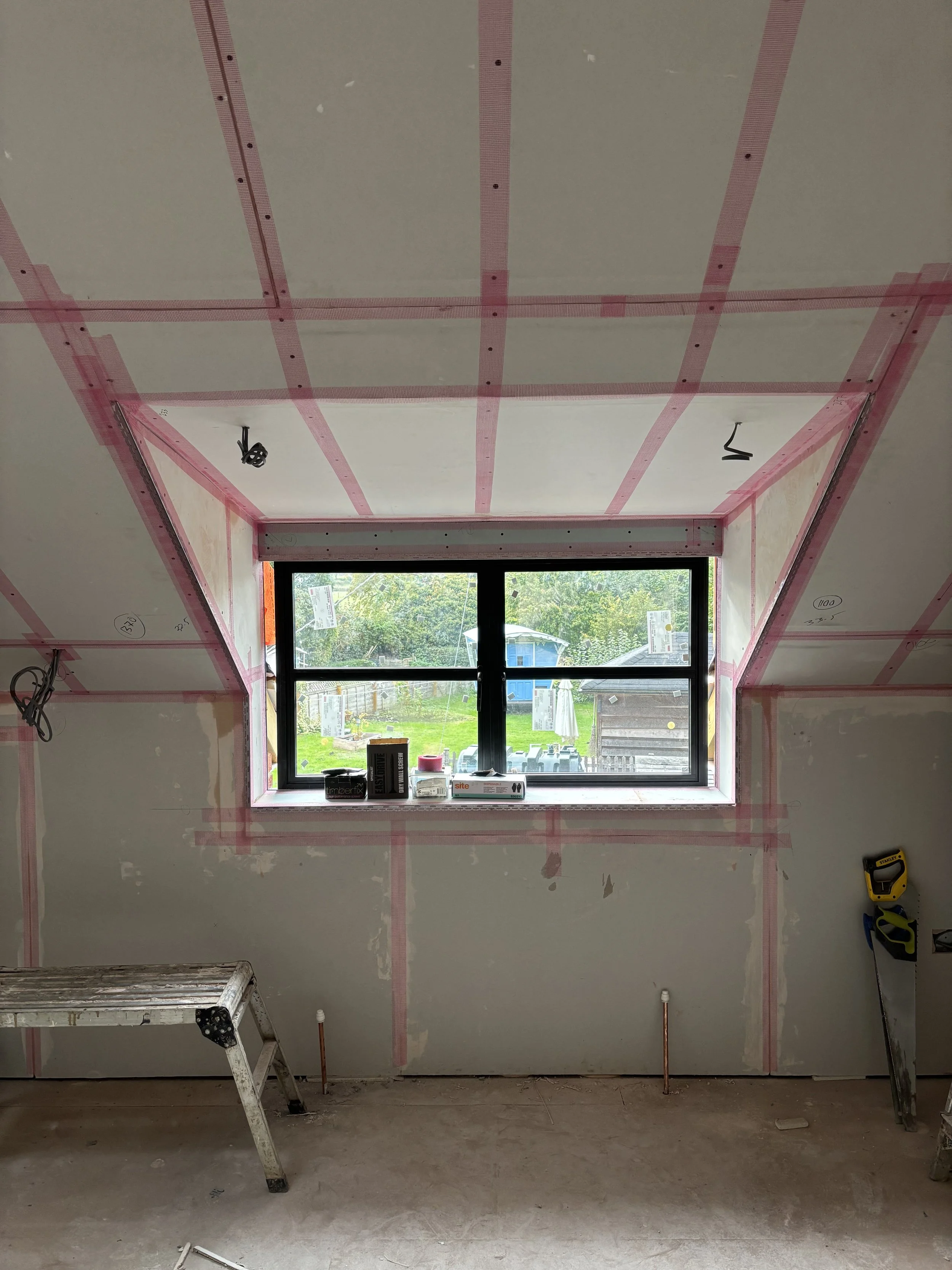 Interior of a room under construction with drywall, window, and tools, showing pink drywall tape and markings on the wall.