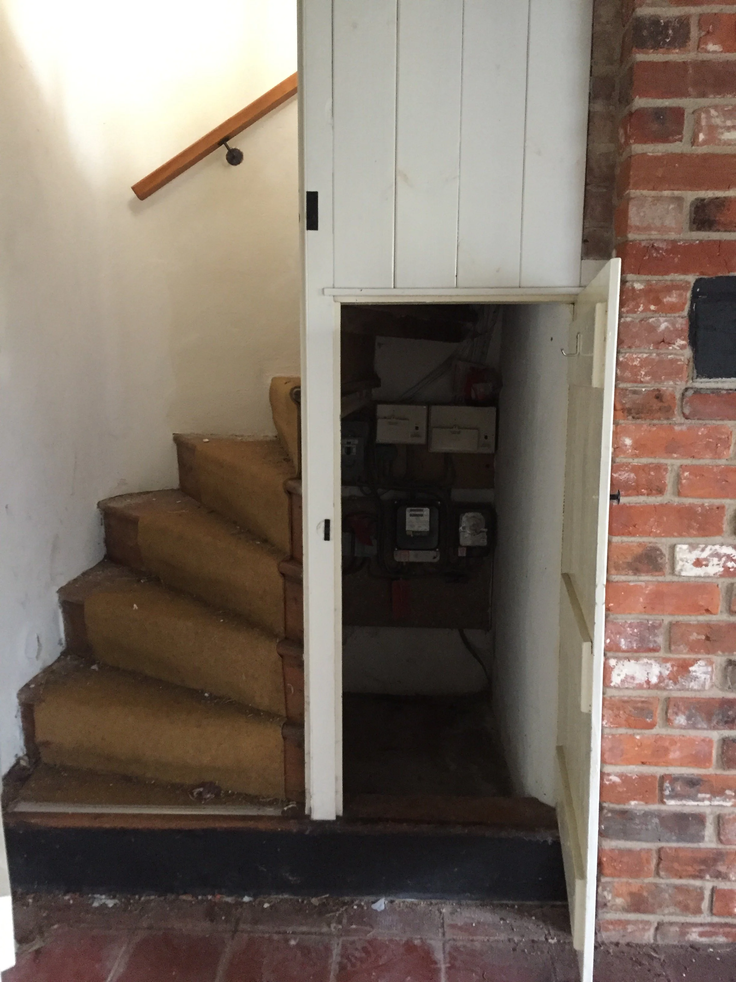 A staircase with three wooden steps leading up to a small white cabinet with a brick wall on the right side. The cabinet is open, revealing some stored items inside. The floor is red tiled, and there is a wall with a wooden handrail on the left side.