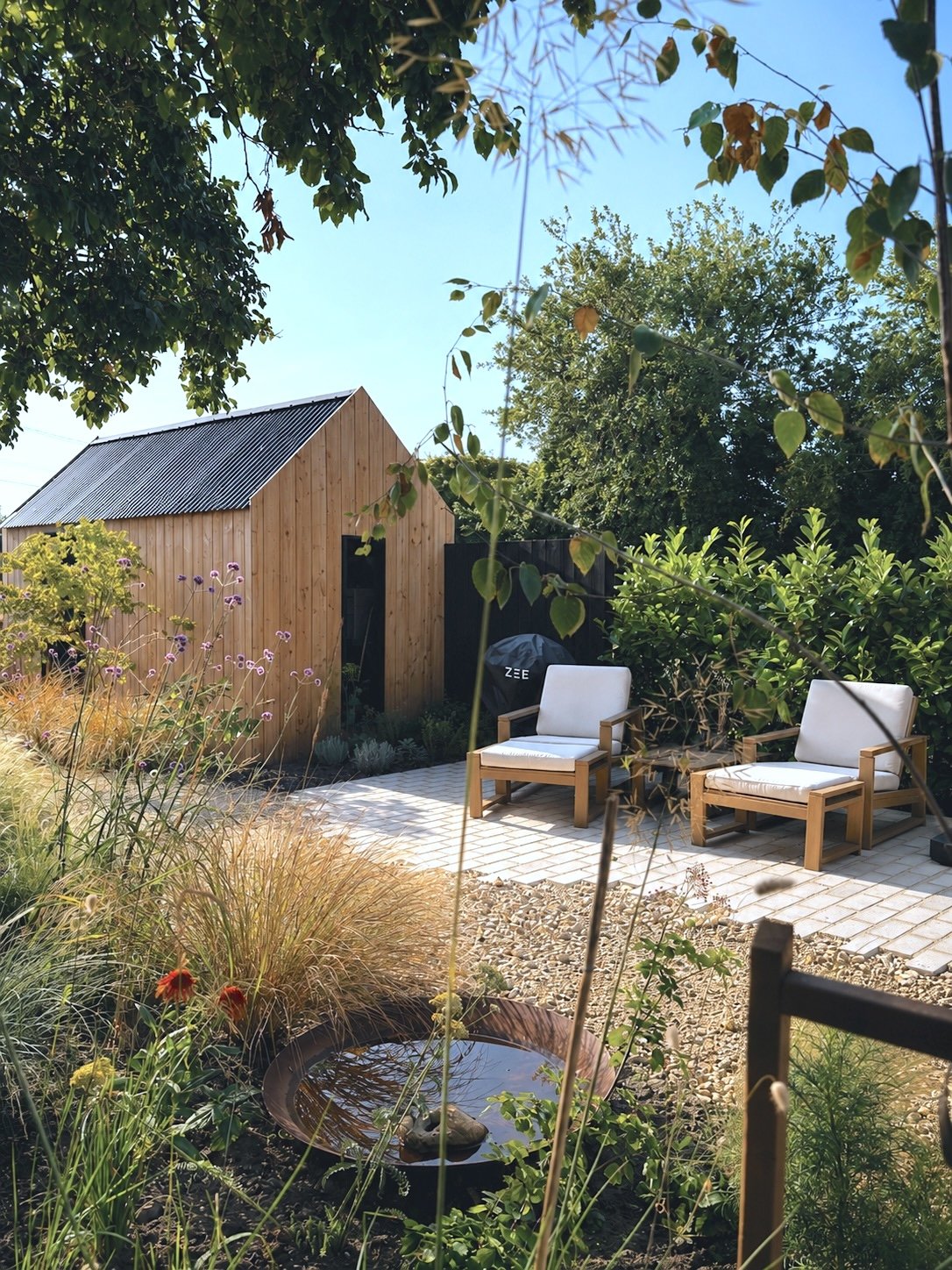 A backyard garden with a wooden shed, two white cushioned chairs, various plants, and a small pond, under a blue sky with some clouds.