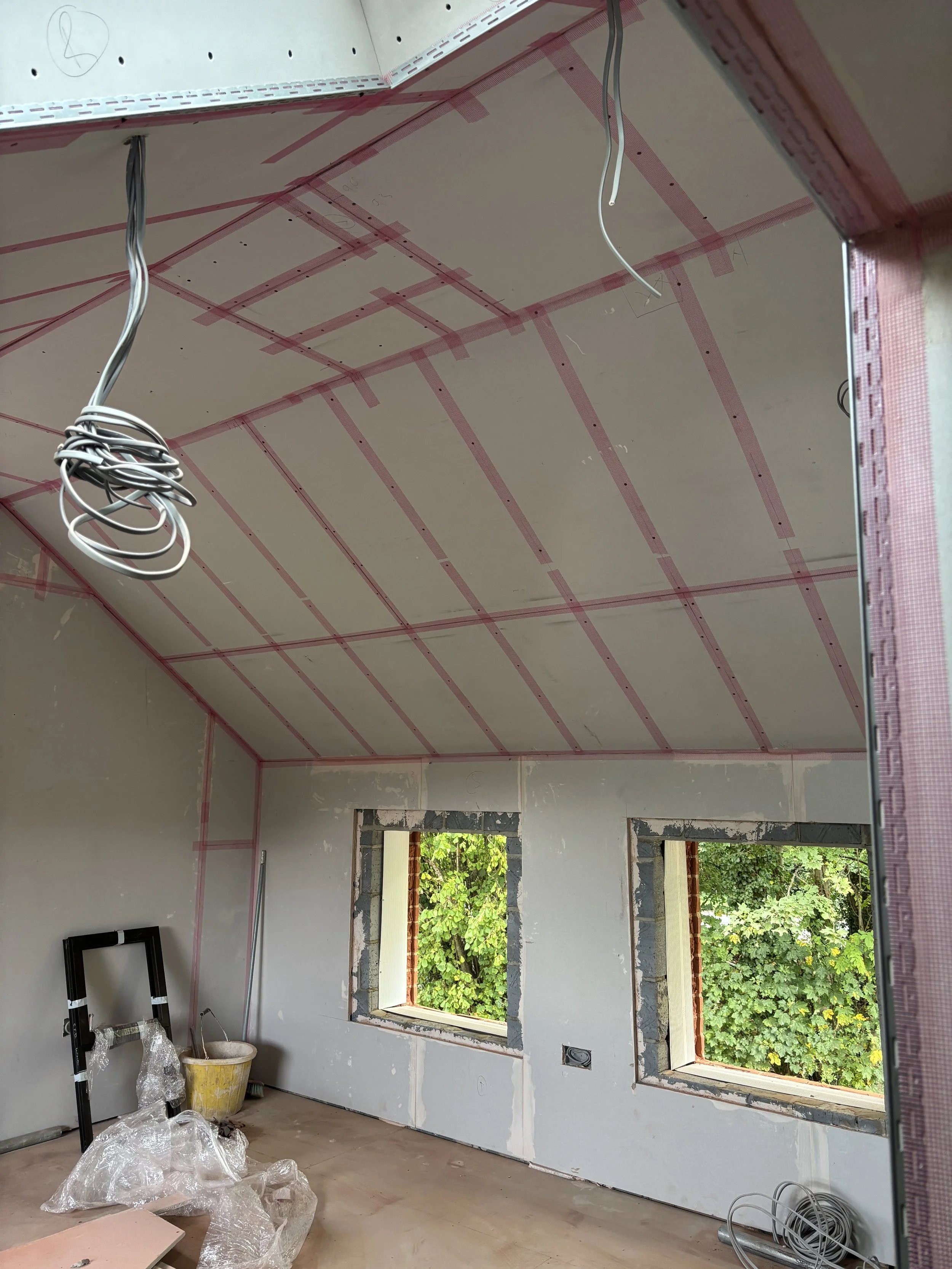 Interior of a room under construction with drywall installed on the ceiling and walls, electrical wiring hanging from the ceiling, and construction materials on the floor.