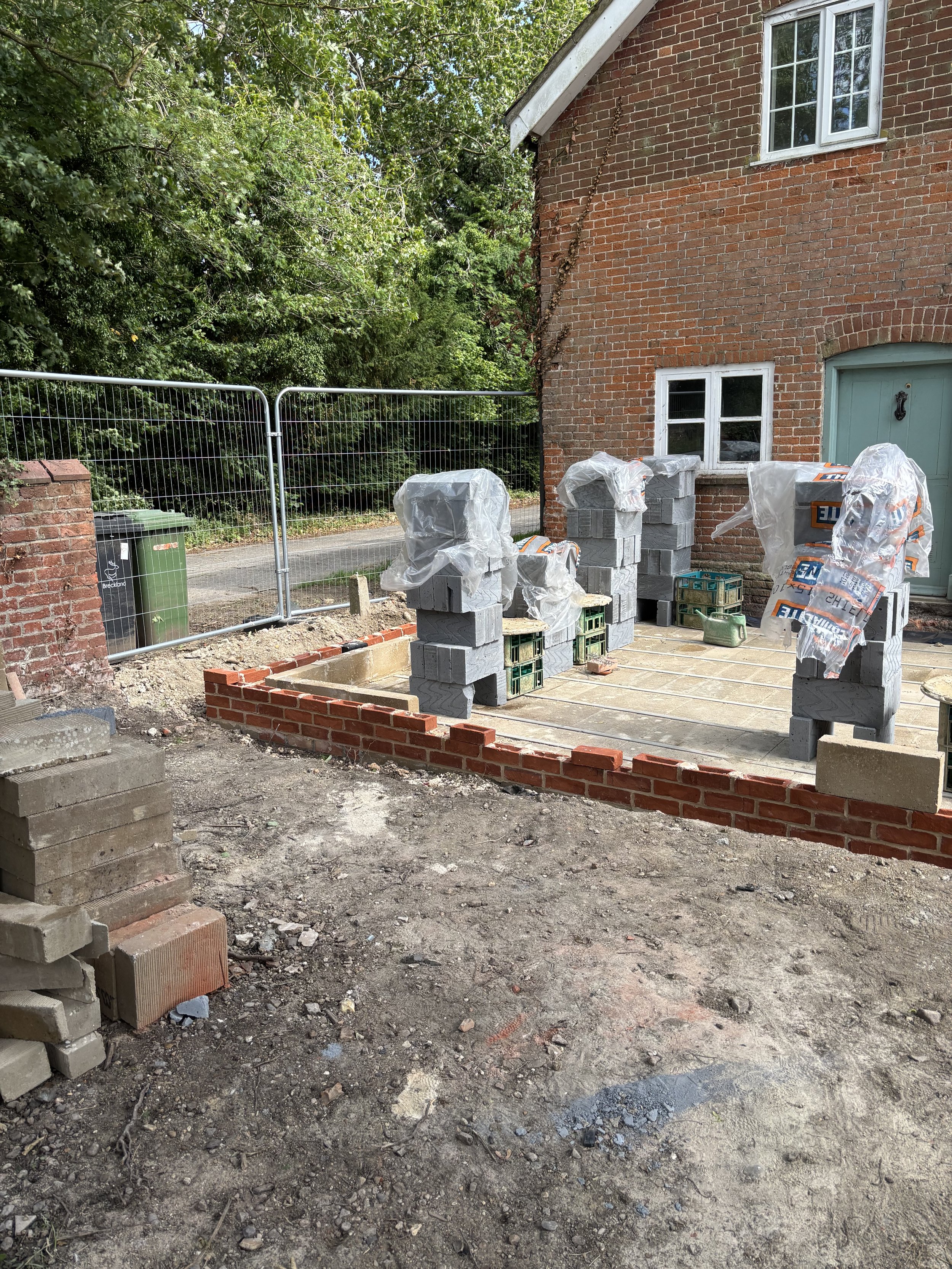 A construction site in front of a brick house with two windows and a green door. Various construction materials, including stacks of bricks and concrete blocks, are scattered around. A temporary metal fence and a green trash bin are also visible. The