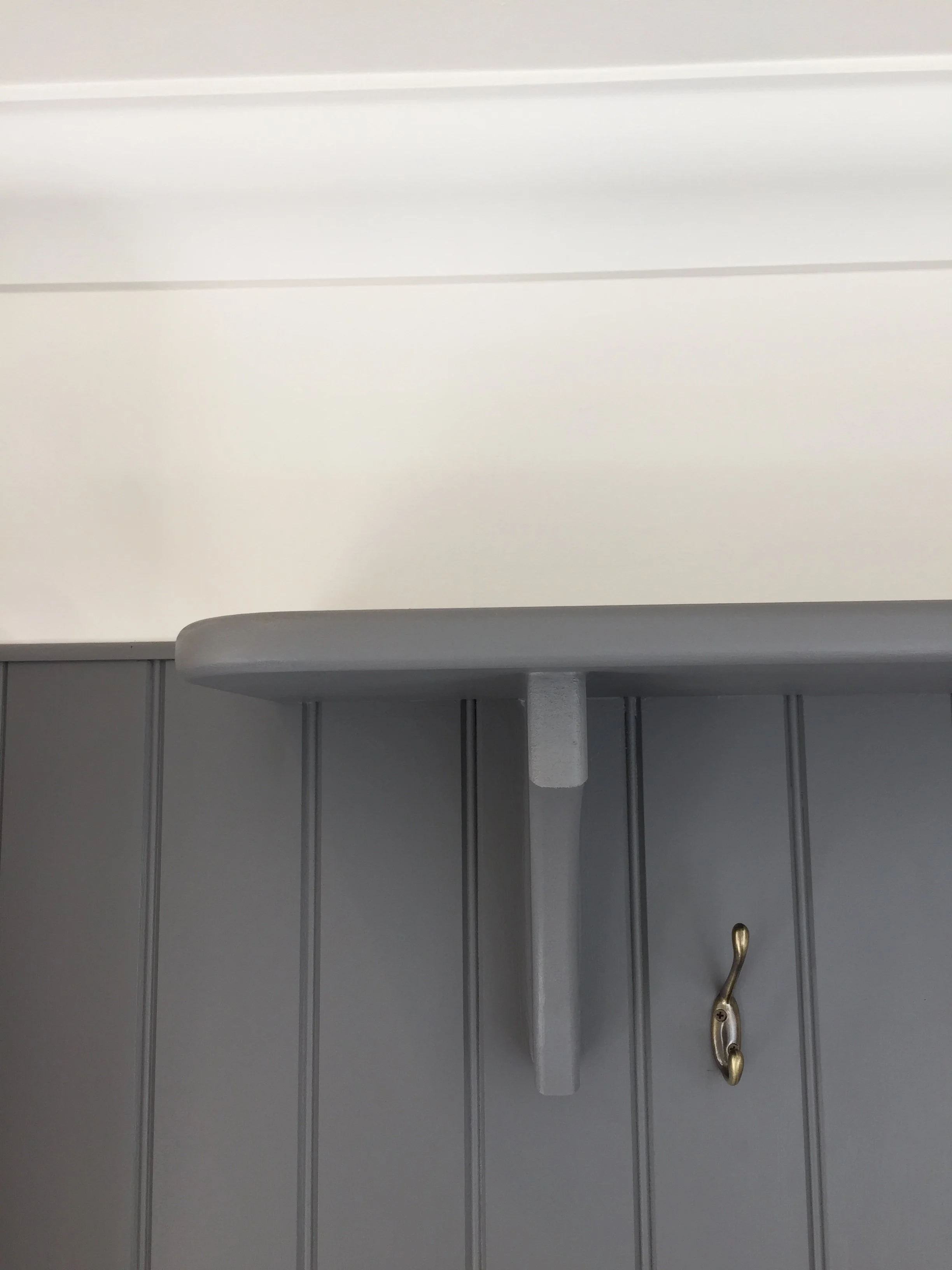 Image of a gray wall with vertical paneling, a gray shelf, and a metallic coat hook with a gold-colored hook.