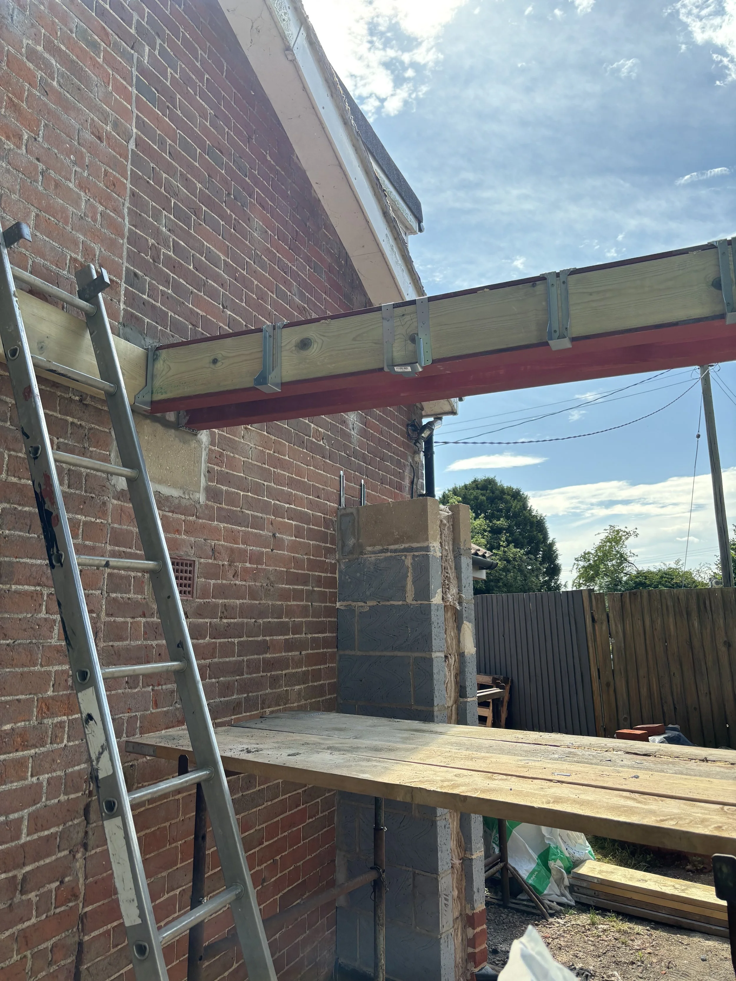 Construction site with a brick building, a ladder, and wooden scaffolding under a partly cloudy sky.