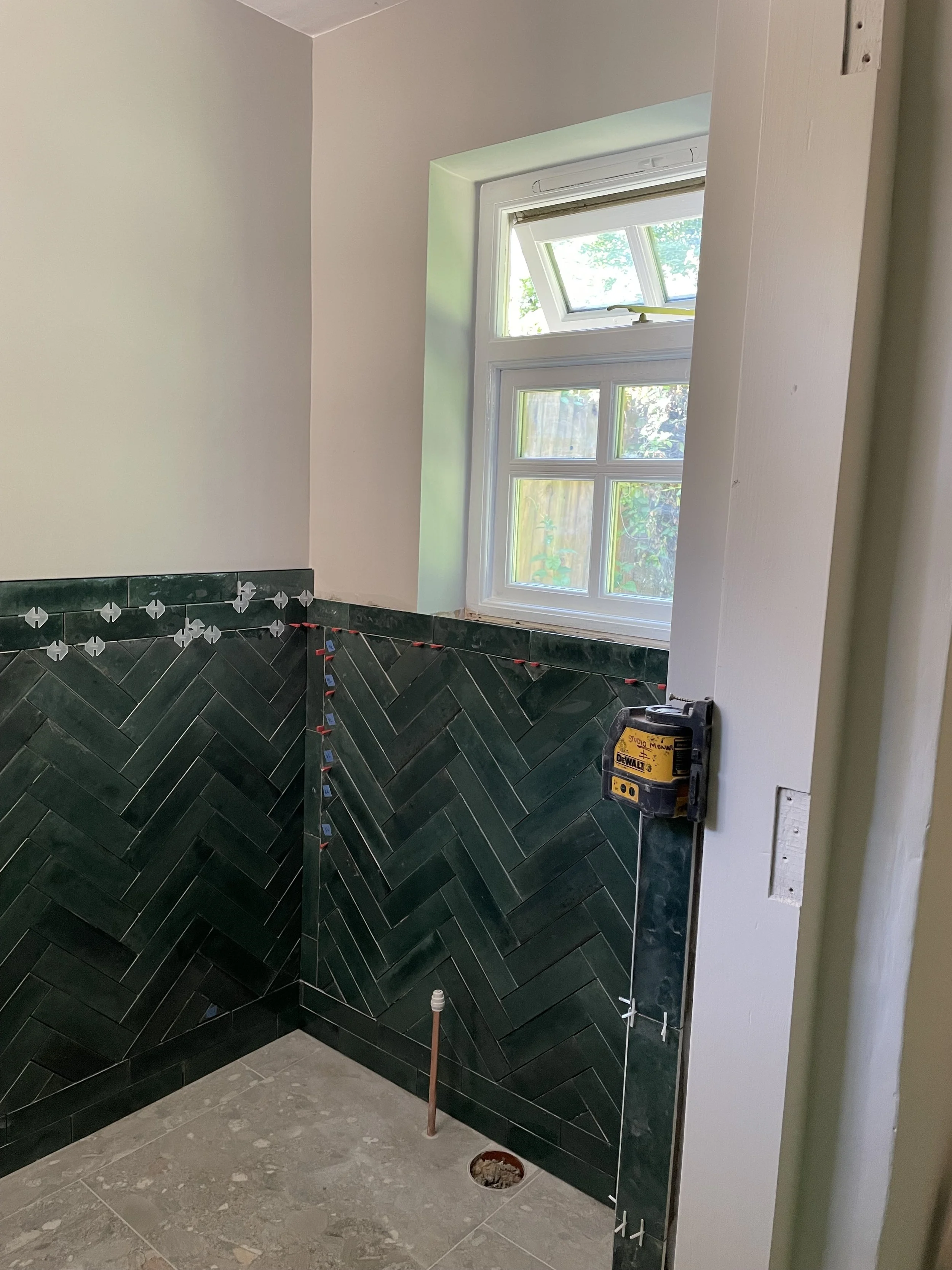 A bathroom under construction with black tile walls, a window, and a pipe sticking out of the floor near the wall. There are tools and construction materials present.