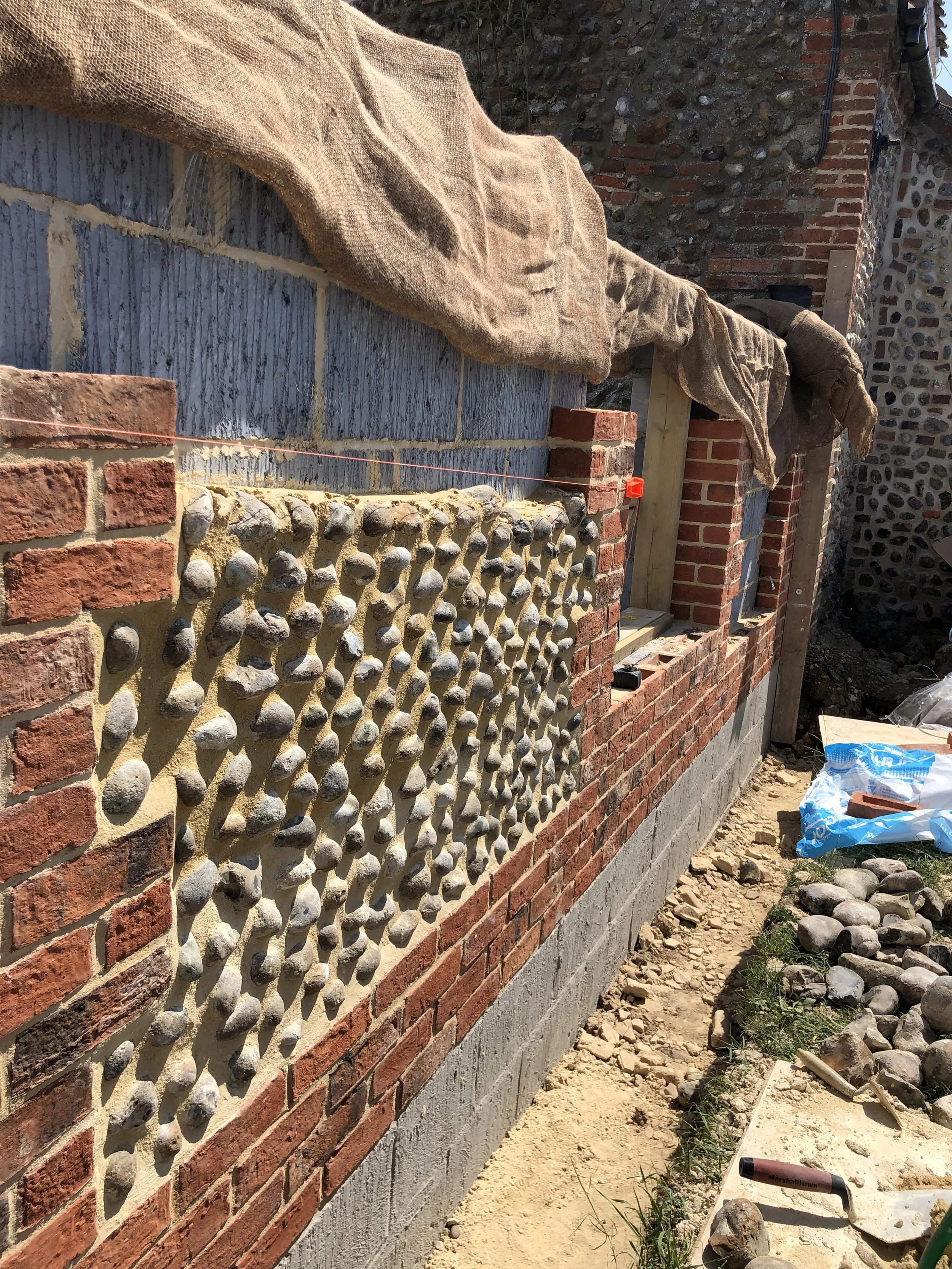 Construction site with brick and stone wall, covered with sandbags and construction tools, and dirt ground with scattered rocks and construction materials.