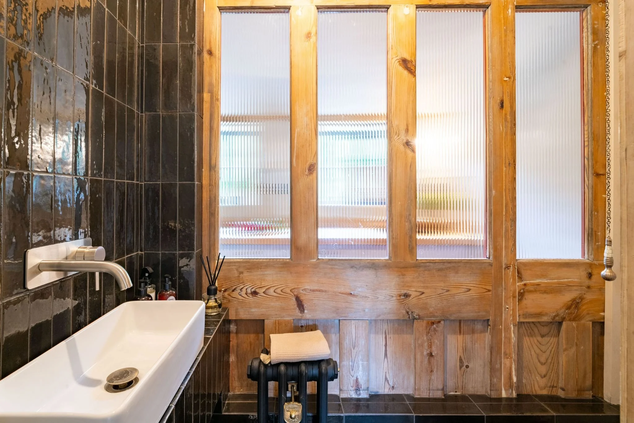 A bathroom with a large window covered by frosted glass and wooden frames. The black tiled wall surrounds a white vessel sink with a modern faucet. On the black tile counter, there are soap dispensers, toiletries, and a reed diffuser. Below the windo