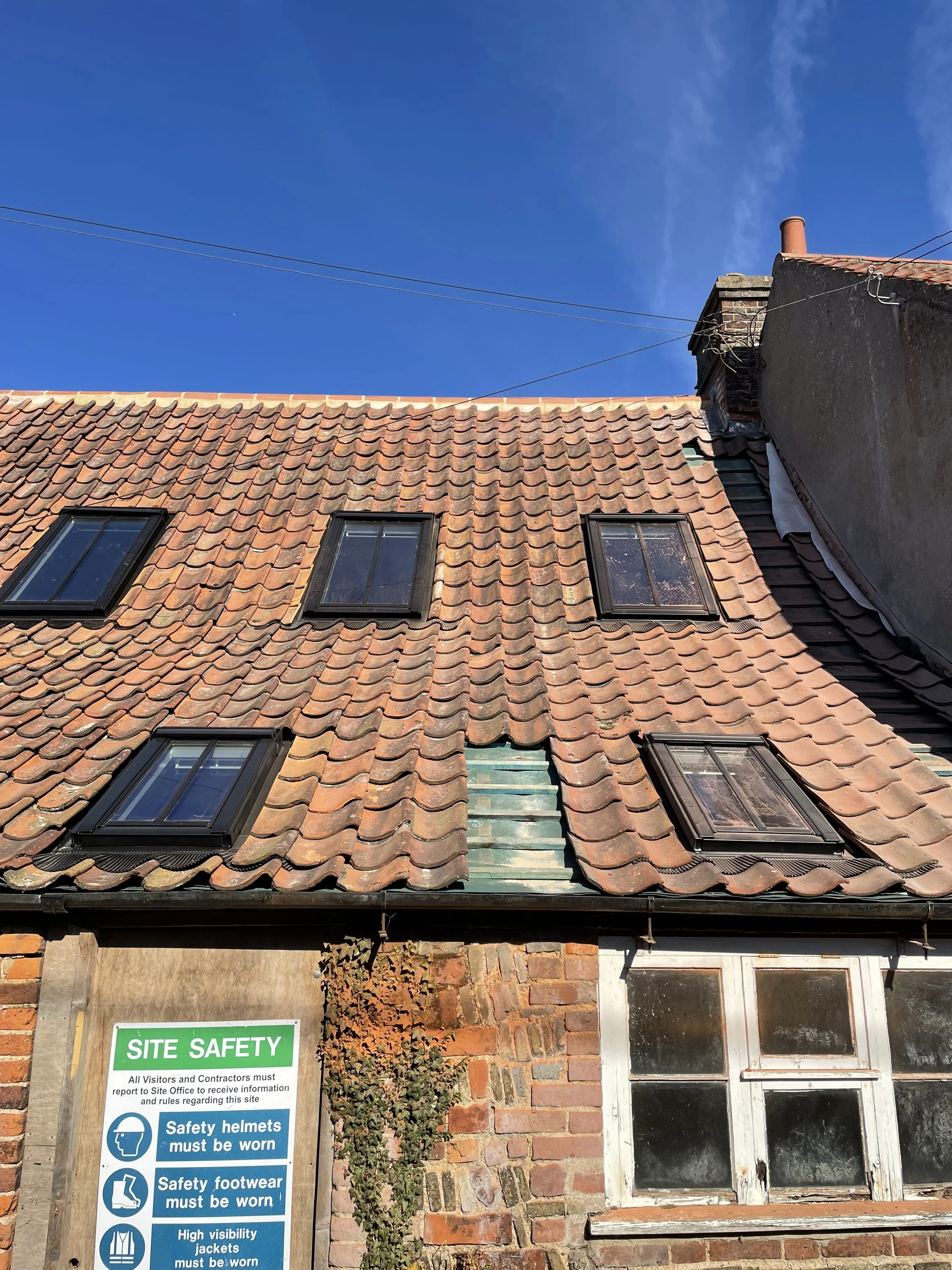 View of a brick building with a sloped tiled roof containing four skylight windows, a chimney, and a sign that says 'Site Safety' with safety instructions, under a clear blue sky.