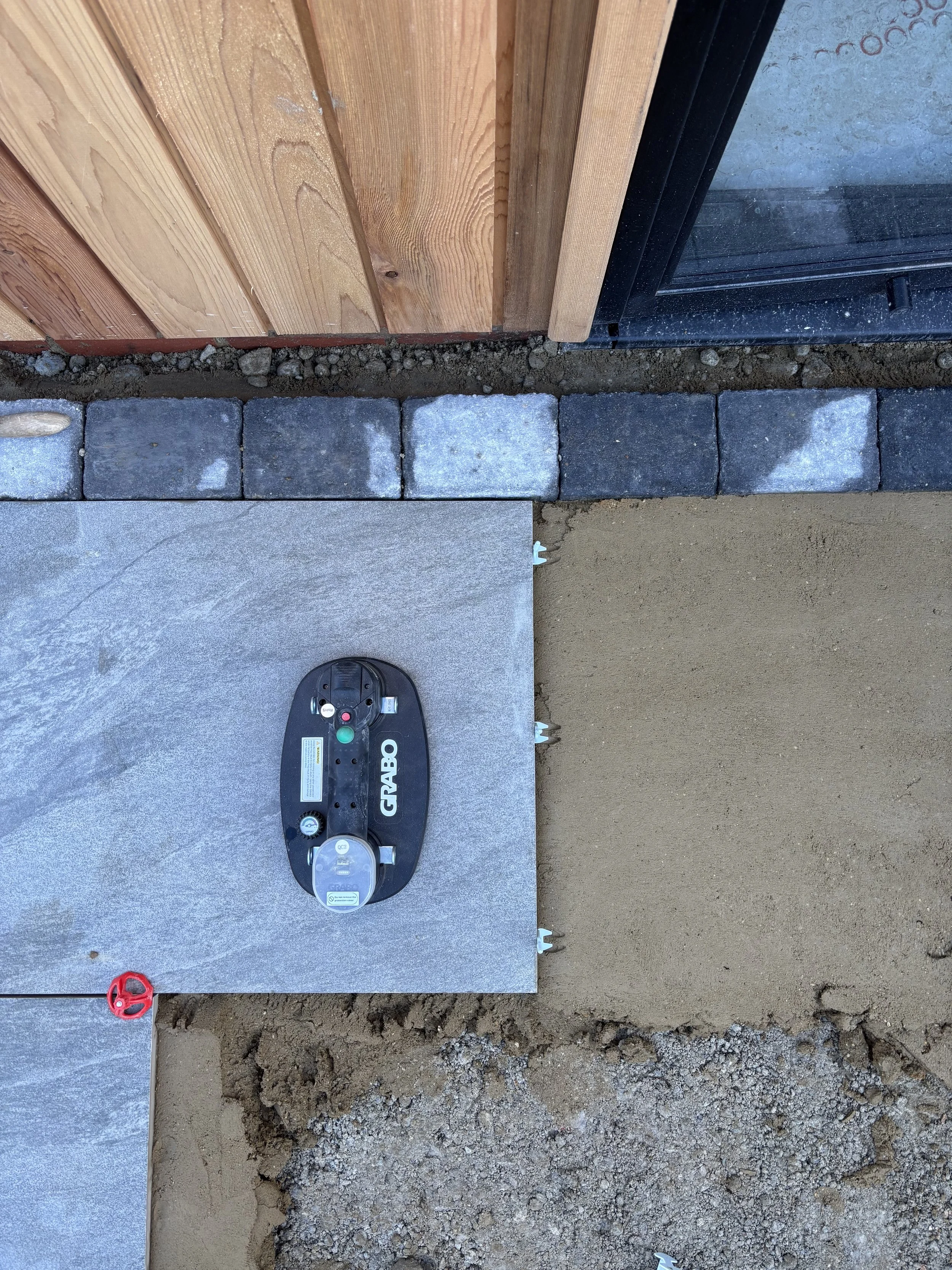 A construction site with a partially installed stone or concrete patio, a plastic temporary cover, a wooden wall on the left, a black window frame on the right, and a red valve in the lower left corner.