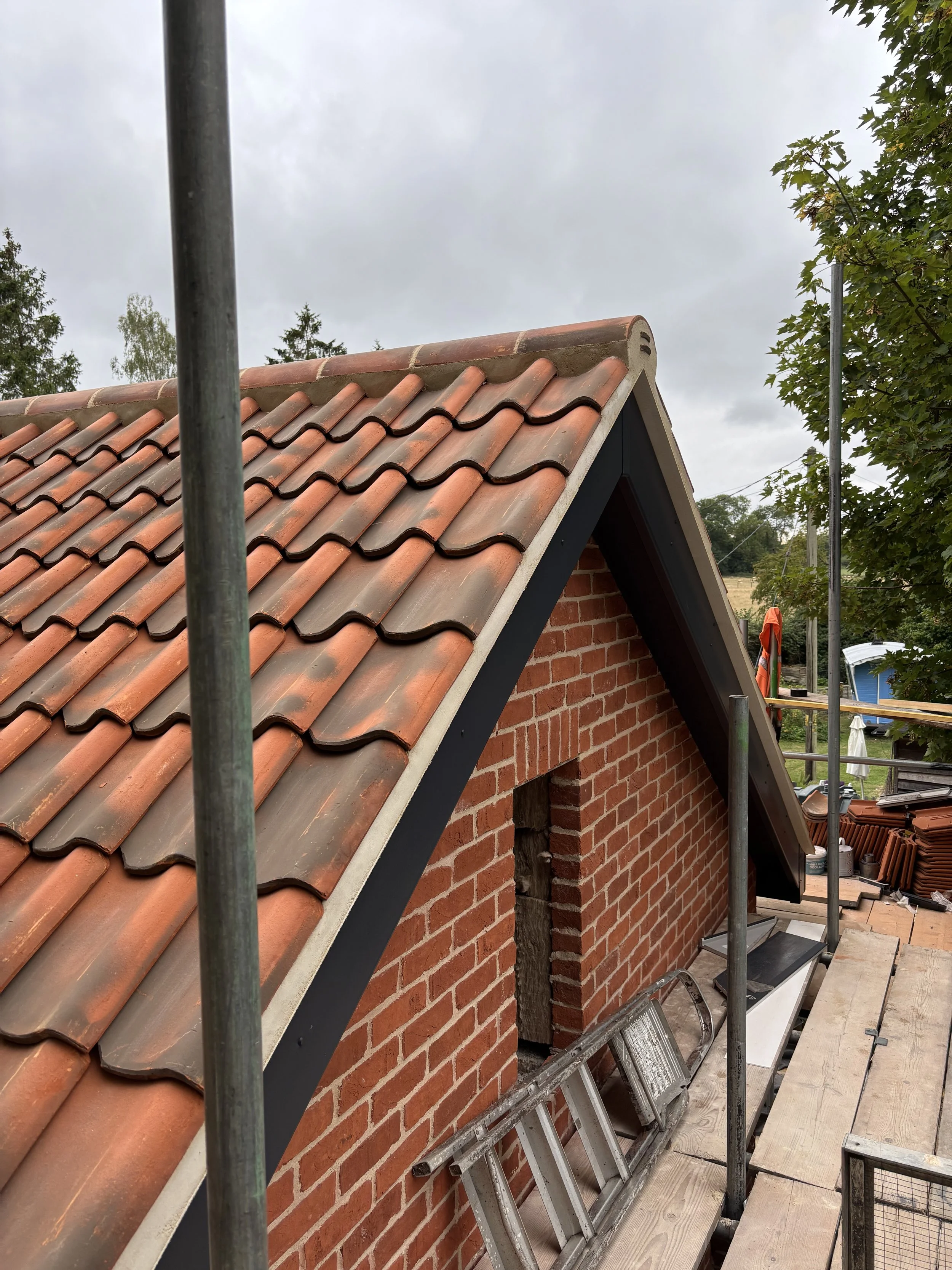 Roof with red clay tiles on a brick building, construction scaffolding and ladder, cloudy sky, and trees in the background.
