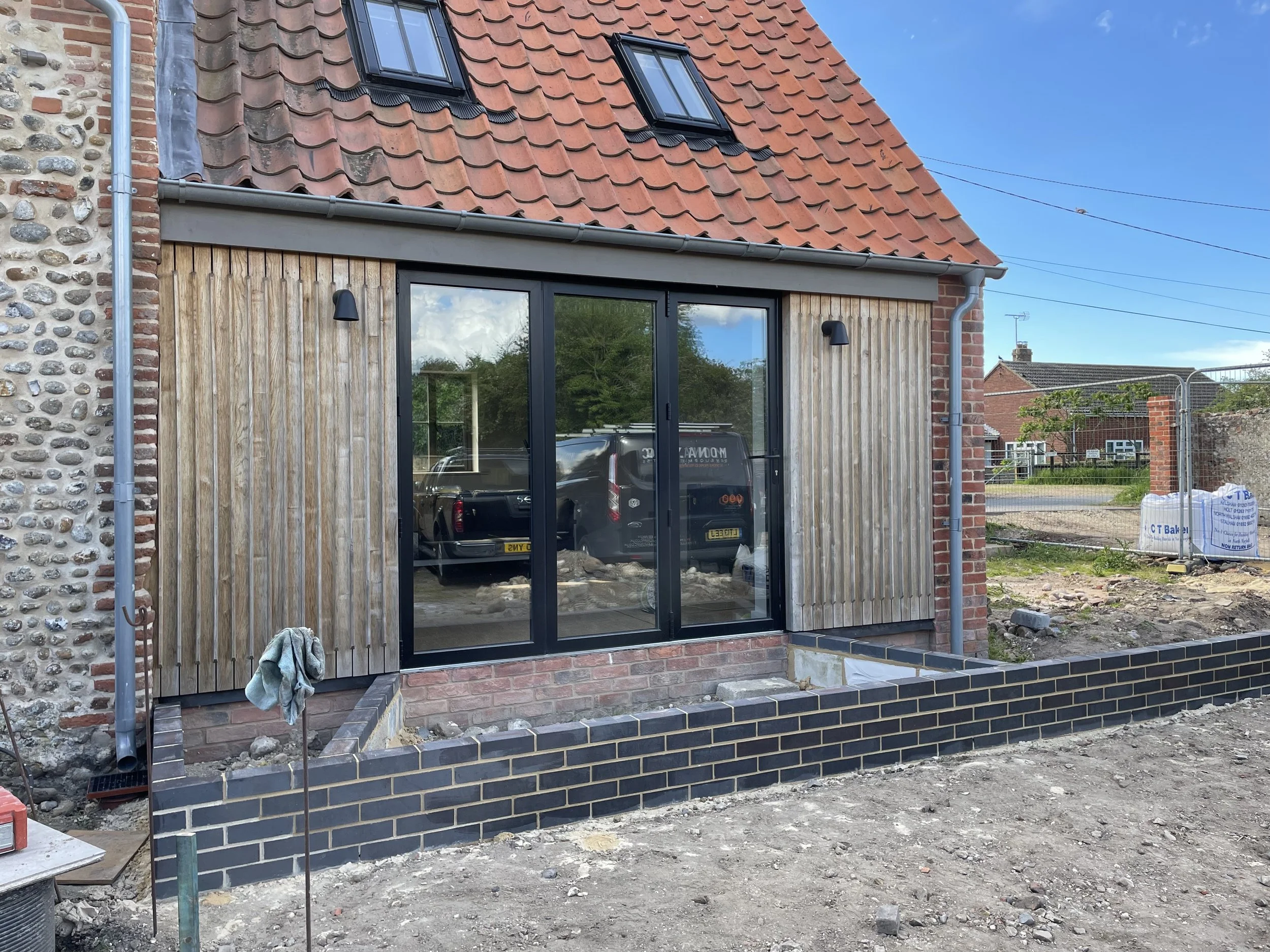 Construction site showing the front of a house with a red tiled roof, black-framed windows, a new wood-paneled wall, and a black brick foundation, with construction materials and equipment on the dirt ground.