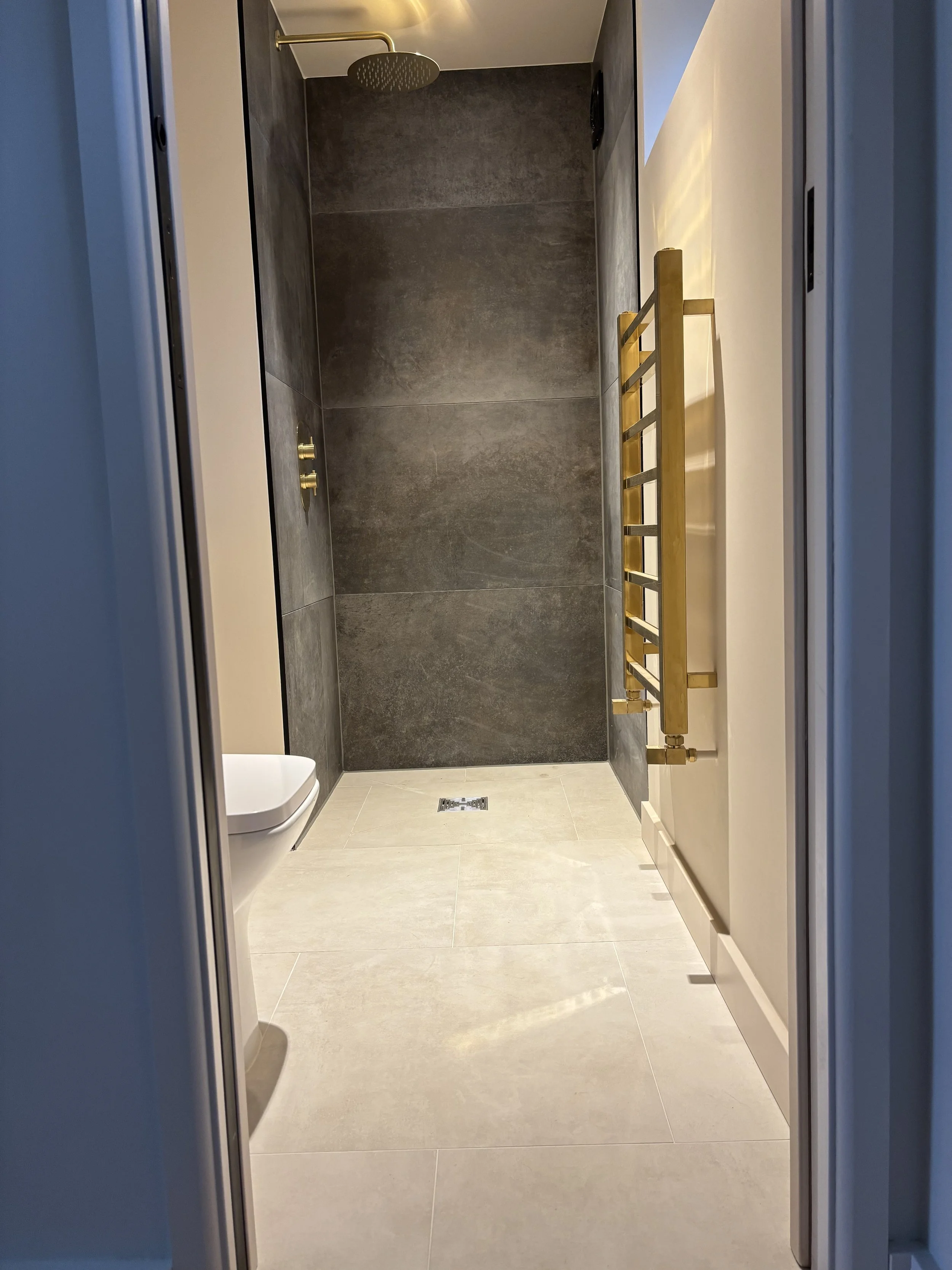 A modern bathroom shower with large dark gray tiles on the wall, a ceiling-mounted rainfall showerhead, gold fixtures, and beige floor tiles.