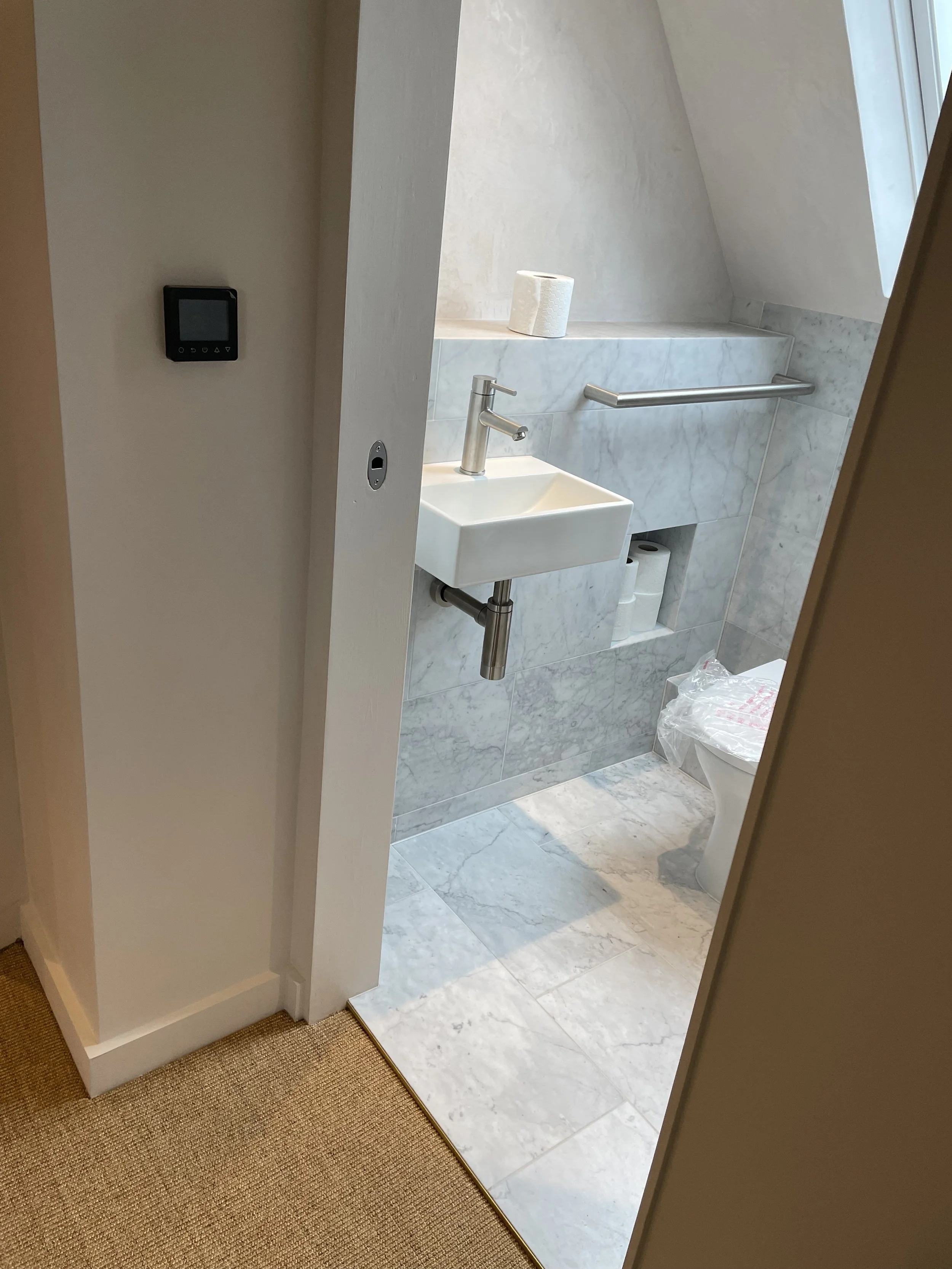 A small half-bathroom with a white square sink, a silver faucet, a paper towel roll, a silver grab bar, and a toilet with a plastic bag on the seat, with marble tiles and a sloped ceiling window.