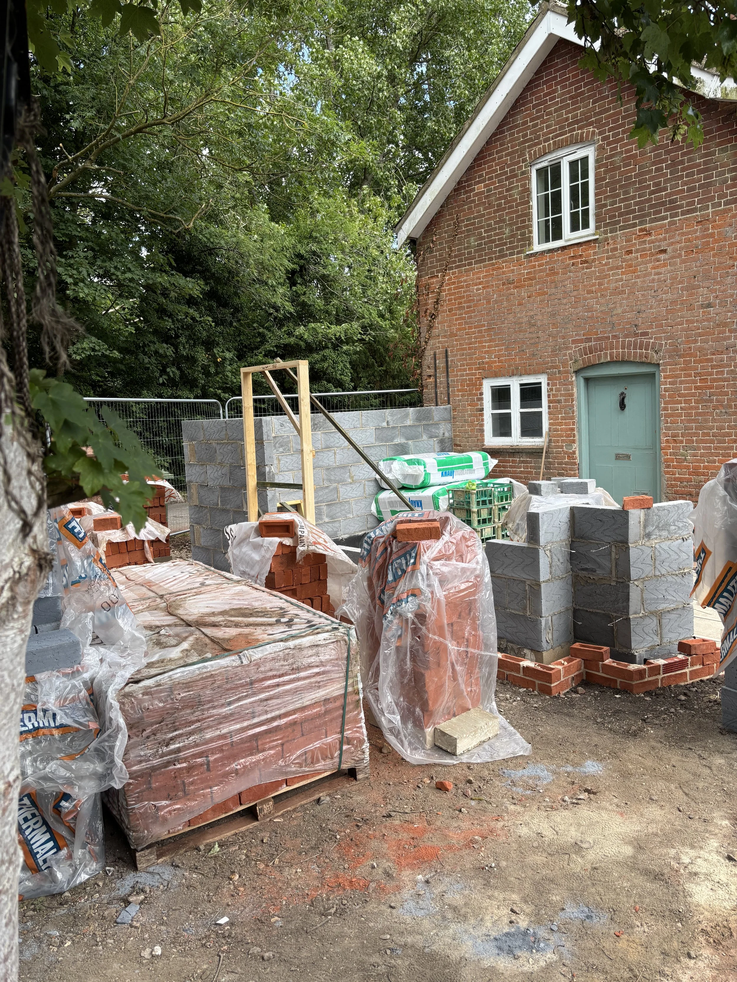 A house under construction with red brick walls, a green door, and white-framed windows, surrounded by construction materials and tools, with trees in the background.