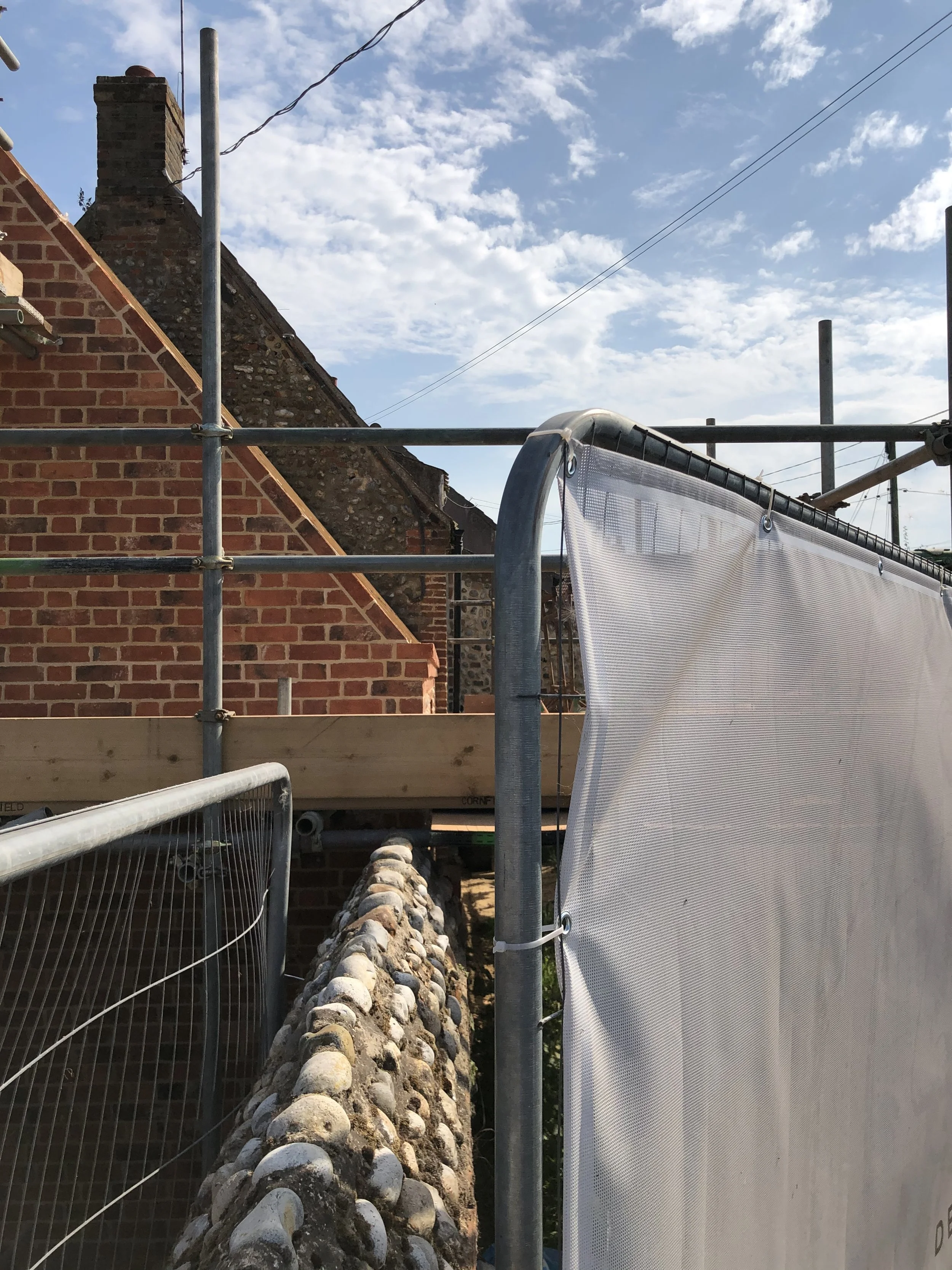 A brick building under construction or renovation surrounded by scaffolding and construction barriers, with a partially visible white construction sheet and an outdoor sky with some clouds.
