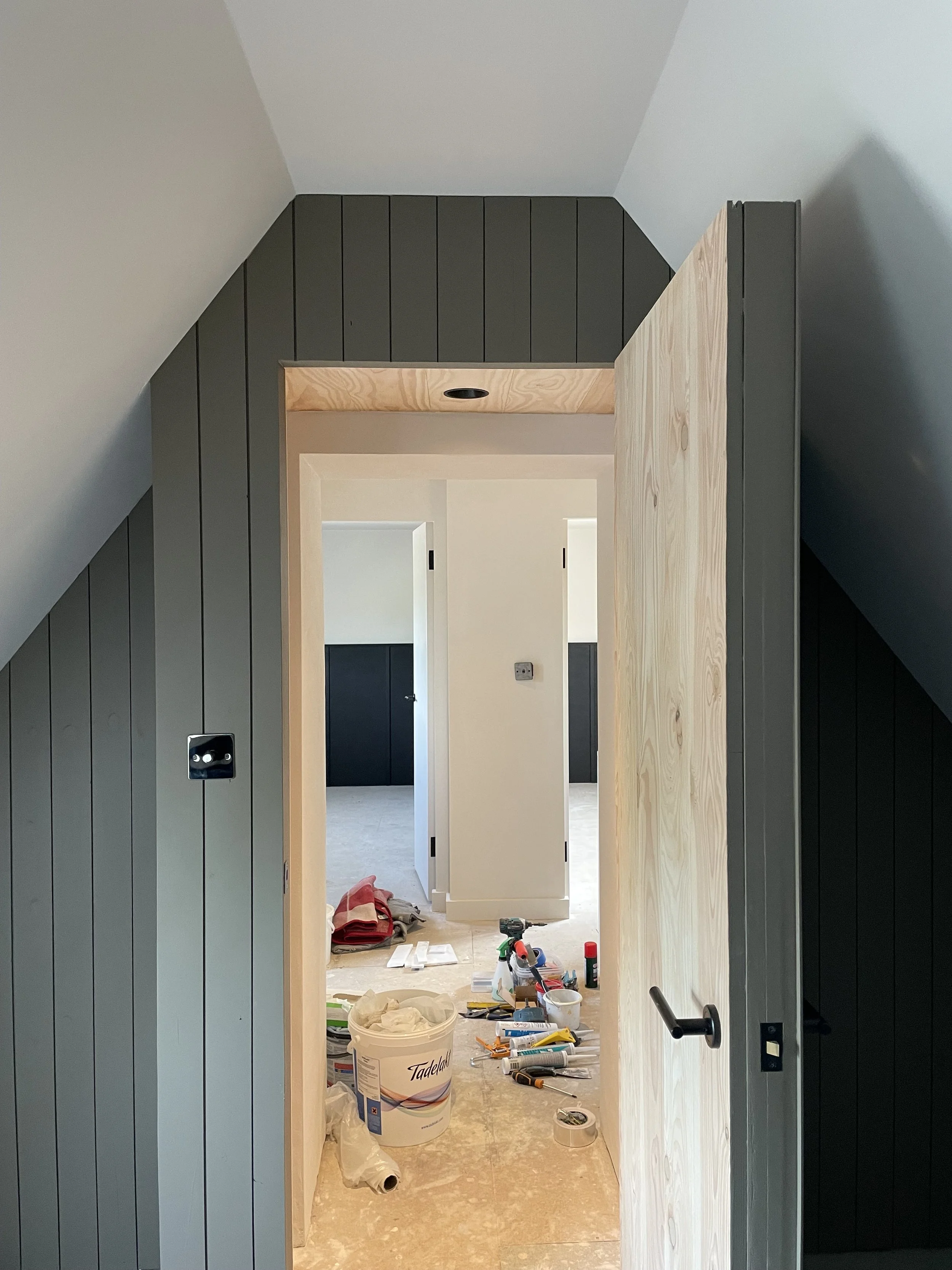 An interior hallway under renovation with tools, paint supplies, and construction materials on the floor, leading to a room with black doors.