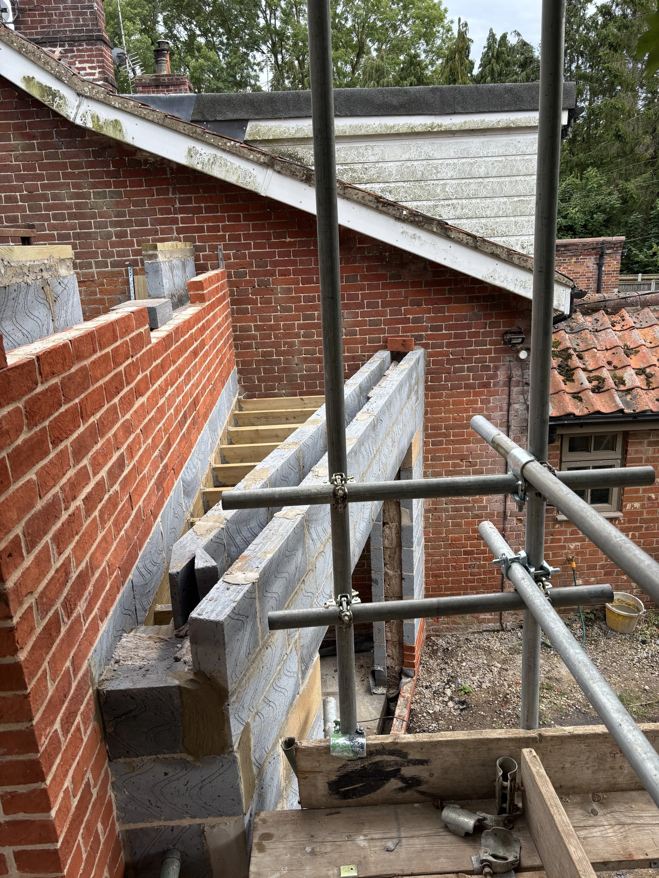 A construction site shows a partially built brick wall and a wooden frame for a new structure. Scaffoldings surround the site, and the background includes trees and a cloudy sky.