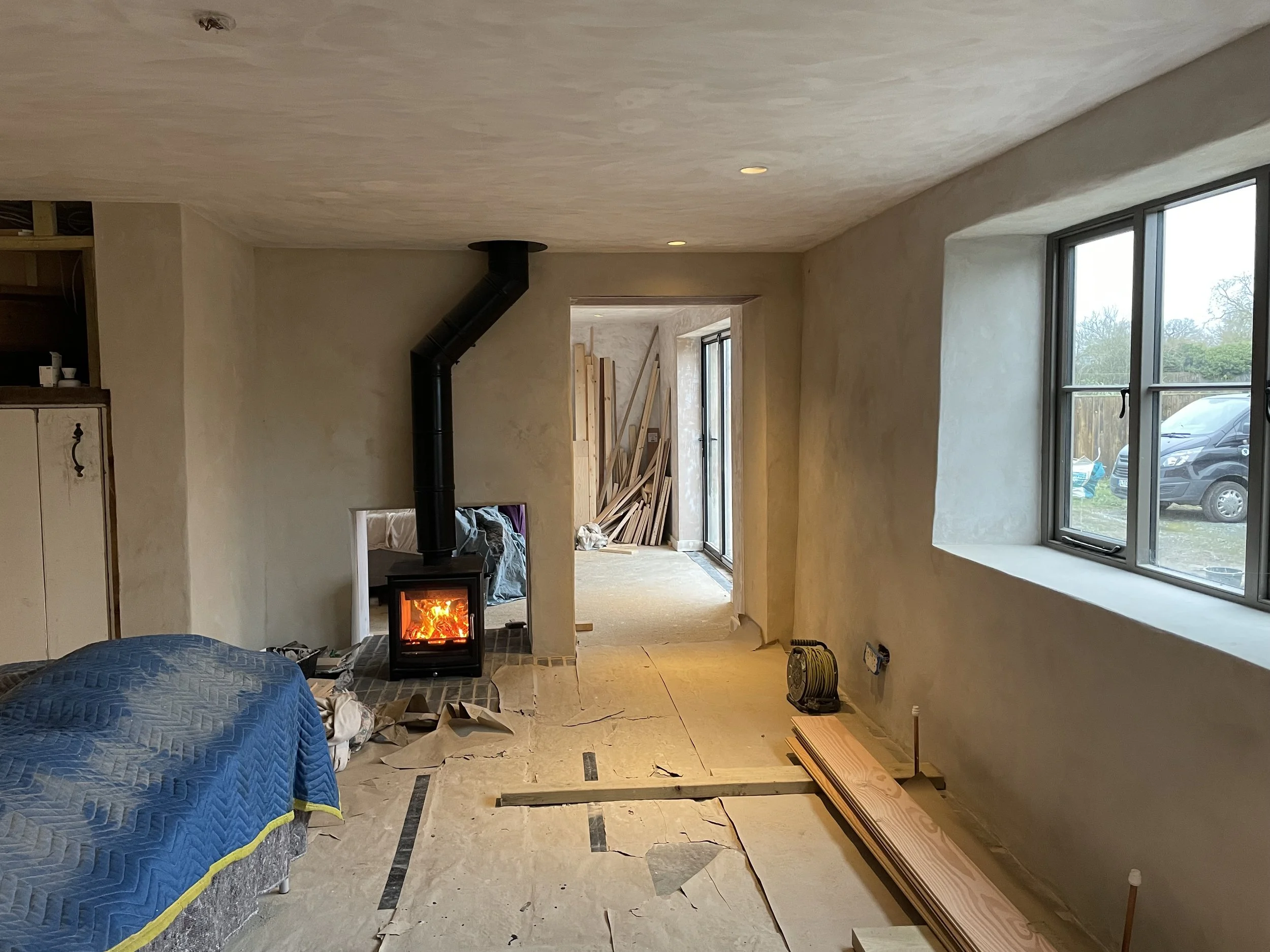 Interior view of a room under renovation with unfinished flooring, a wood stove, and construction materials. A window shows a parked car outside on a cloudy day.