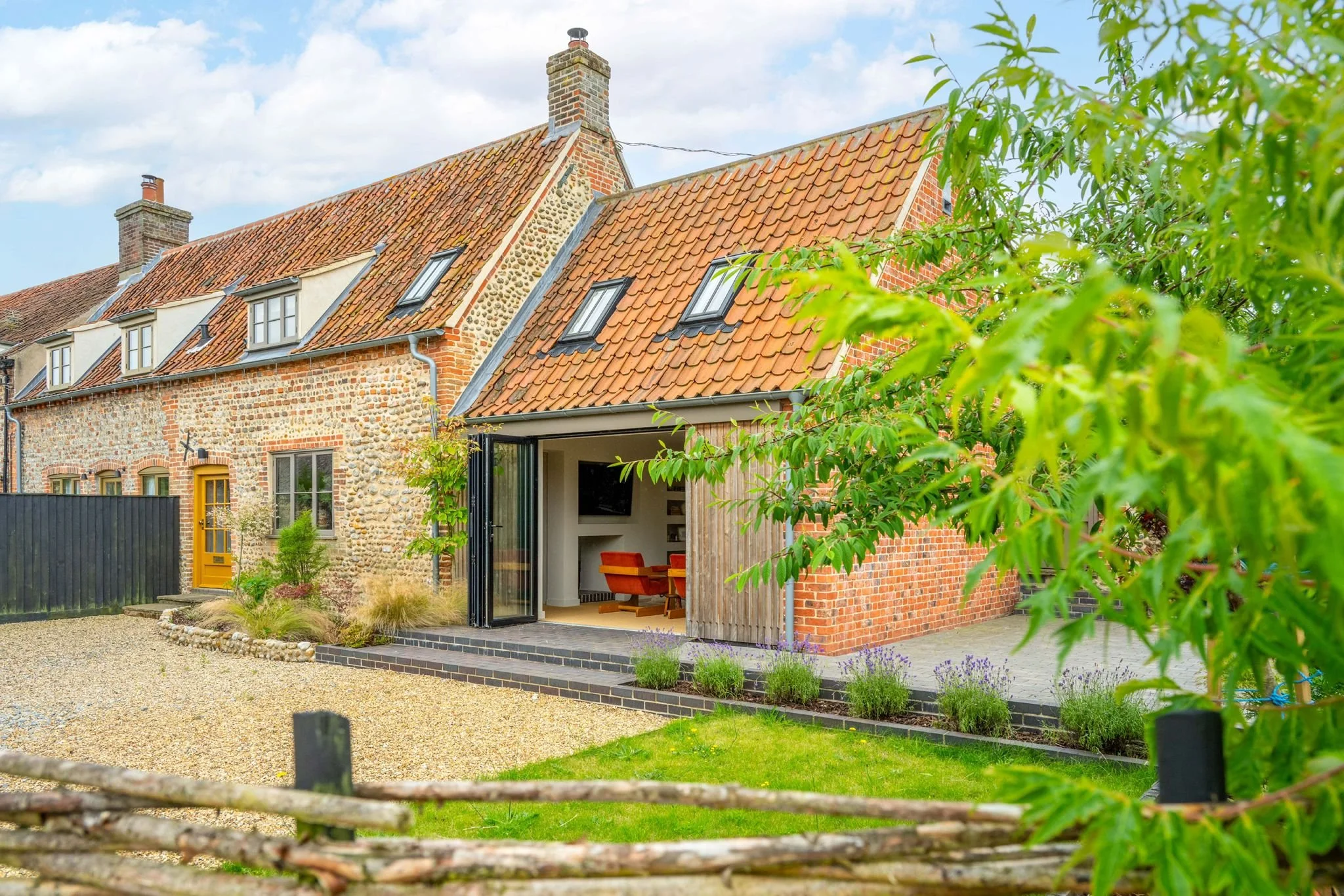 A house with a brick and stone facade, red tiled roof, and multiple small dormer windows. The house features a modern extension with large sliding glass doors opening to a patio and garden, and has a small backyard with plants and a gravel pathway.