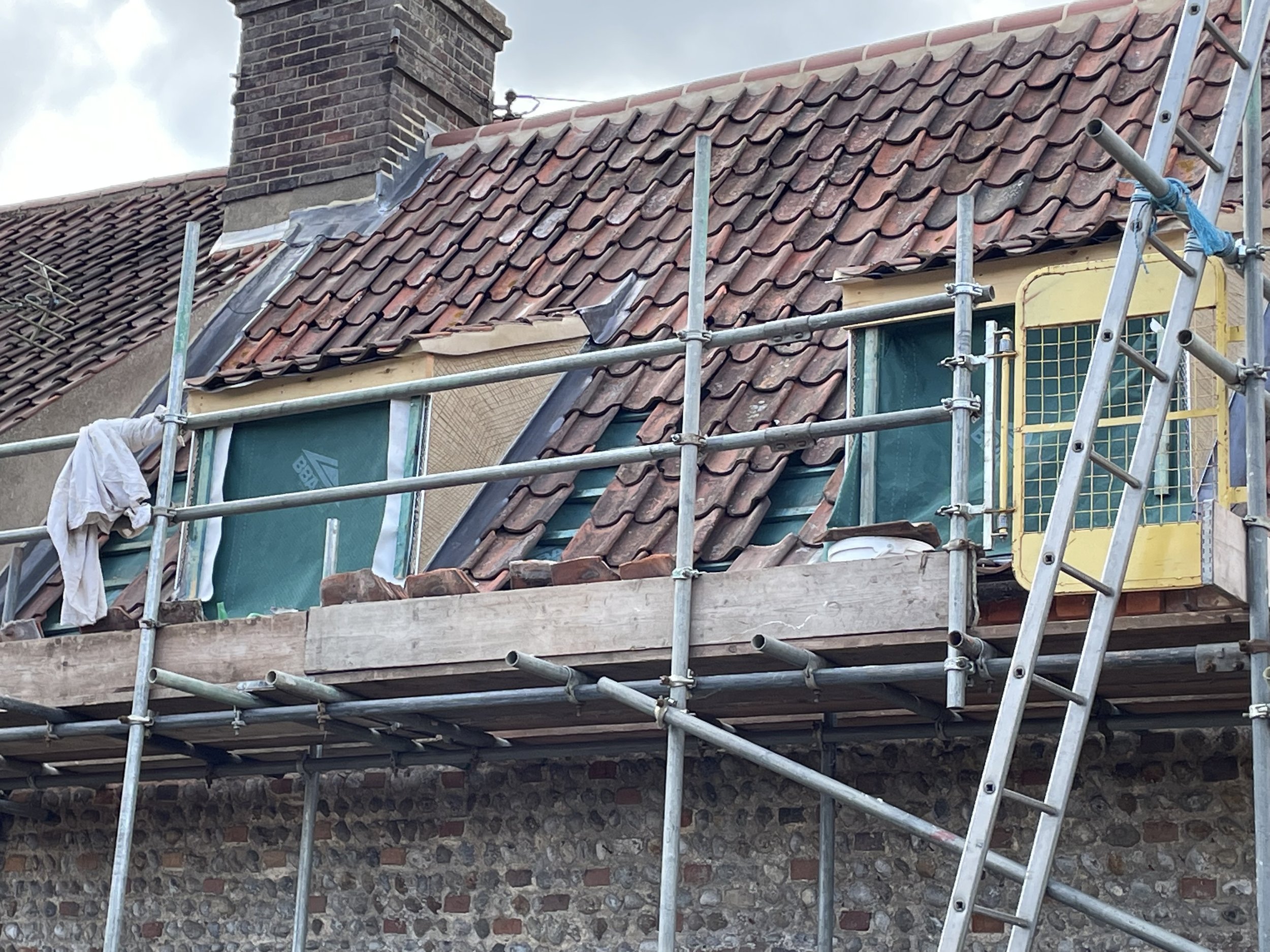Scaffolding set up around a house with a roof being repaired or renovated, showing roof tiles, chimney, and construction materials.